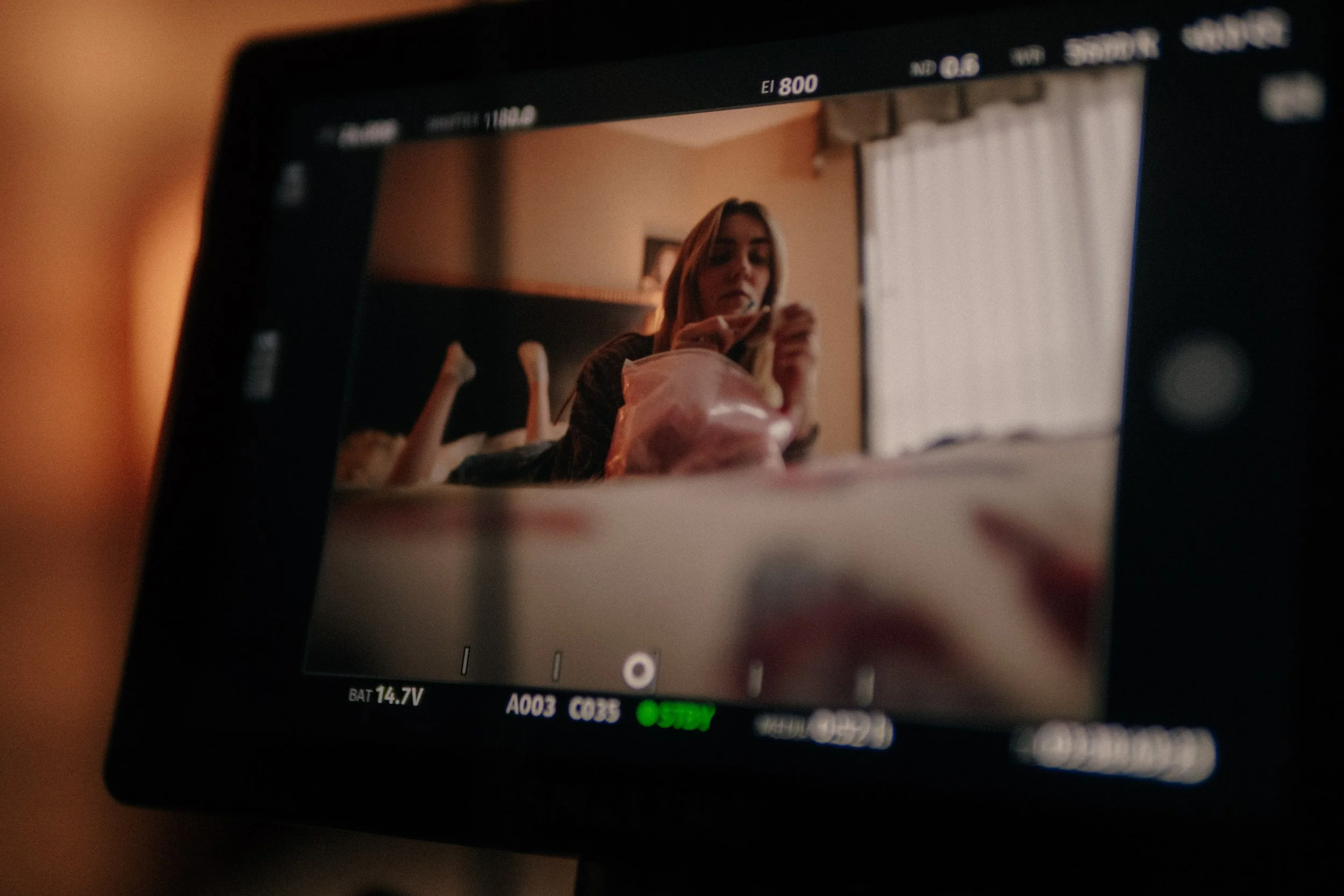 A camera screen showing a young woman sitting on a bed, looking at her phone, with feet raised and crossed in the background, in a room with a window and curtains.