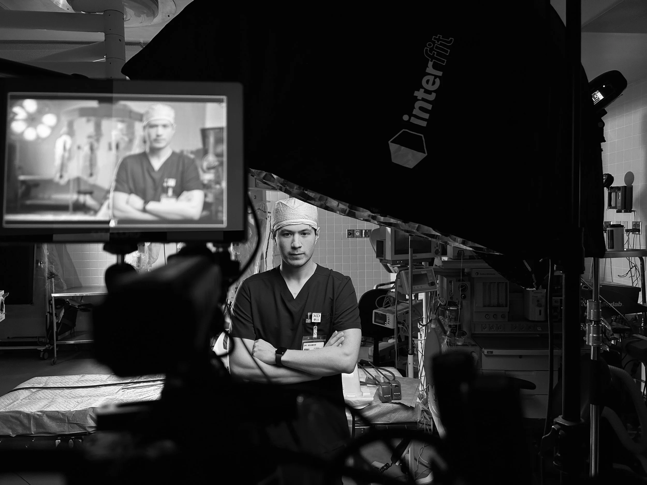 Black and white photograph of a male doctor or surgeon in scrubs and a surgical cap, standing with arms crossed in a hospital or operating room, with a camera and filming equipment in the foreground capturing his image.