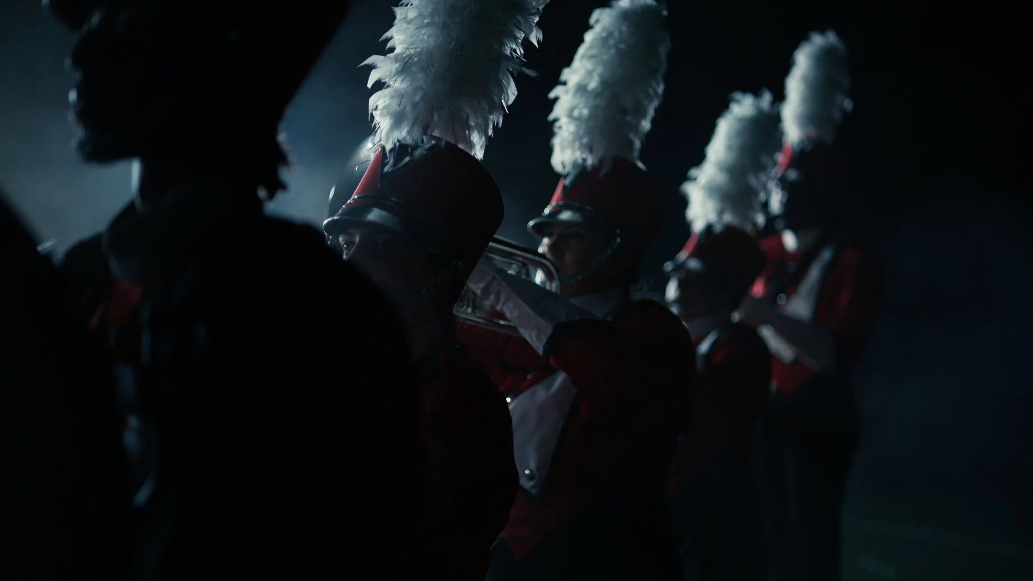 Marching band members in uniform, wearing tall feathered hats, preparing for a performance in a dark setting.