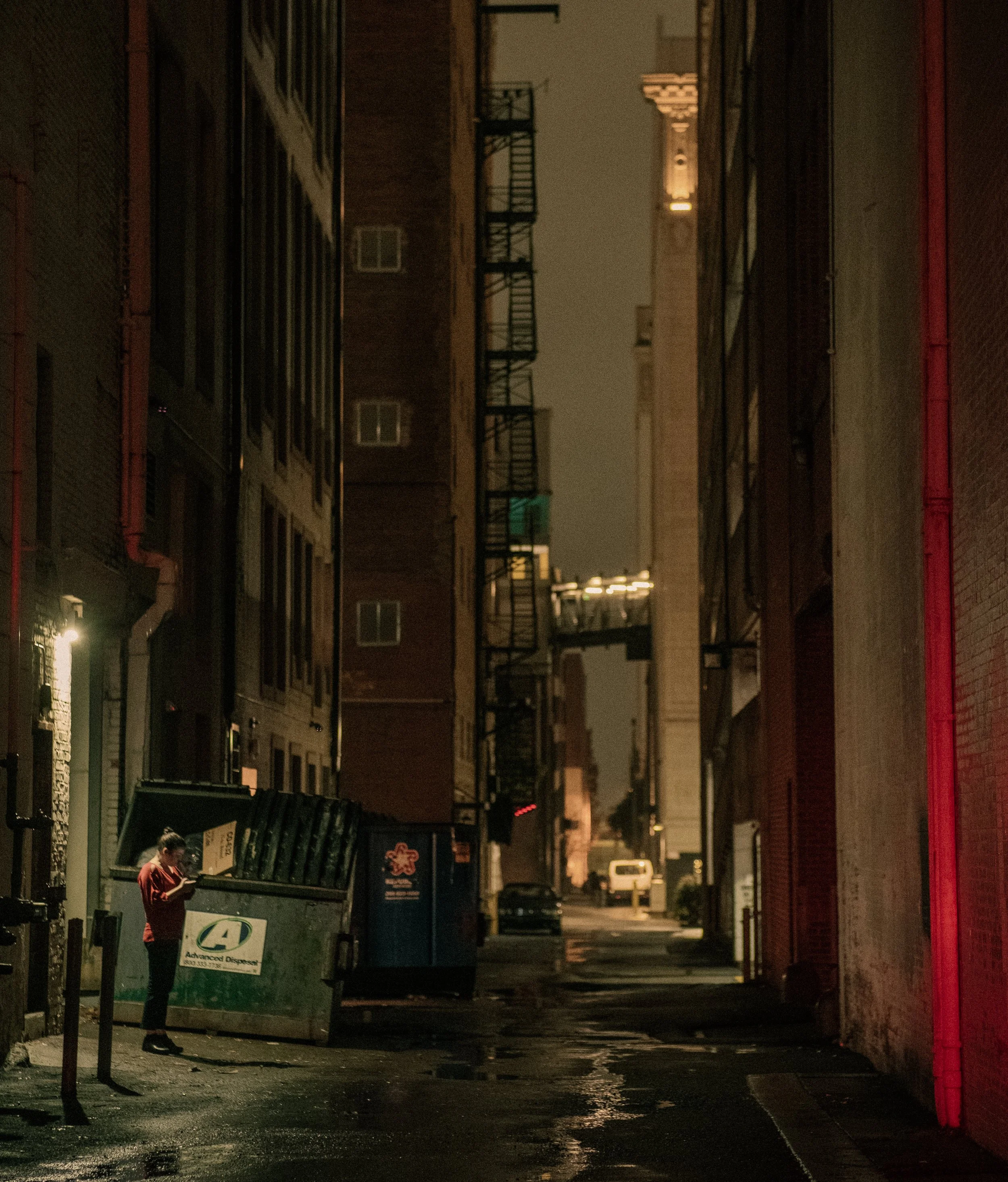A person standing next to a dumpster in a dark, urban alley at night.