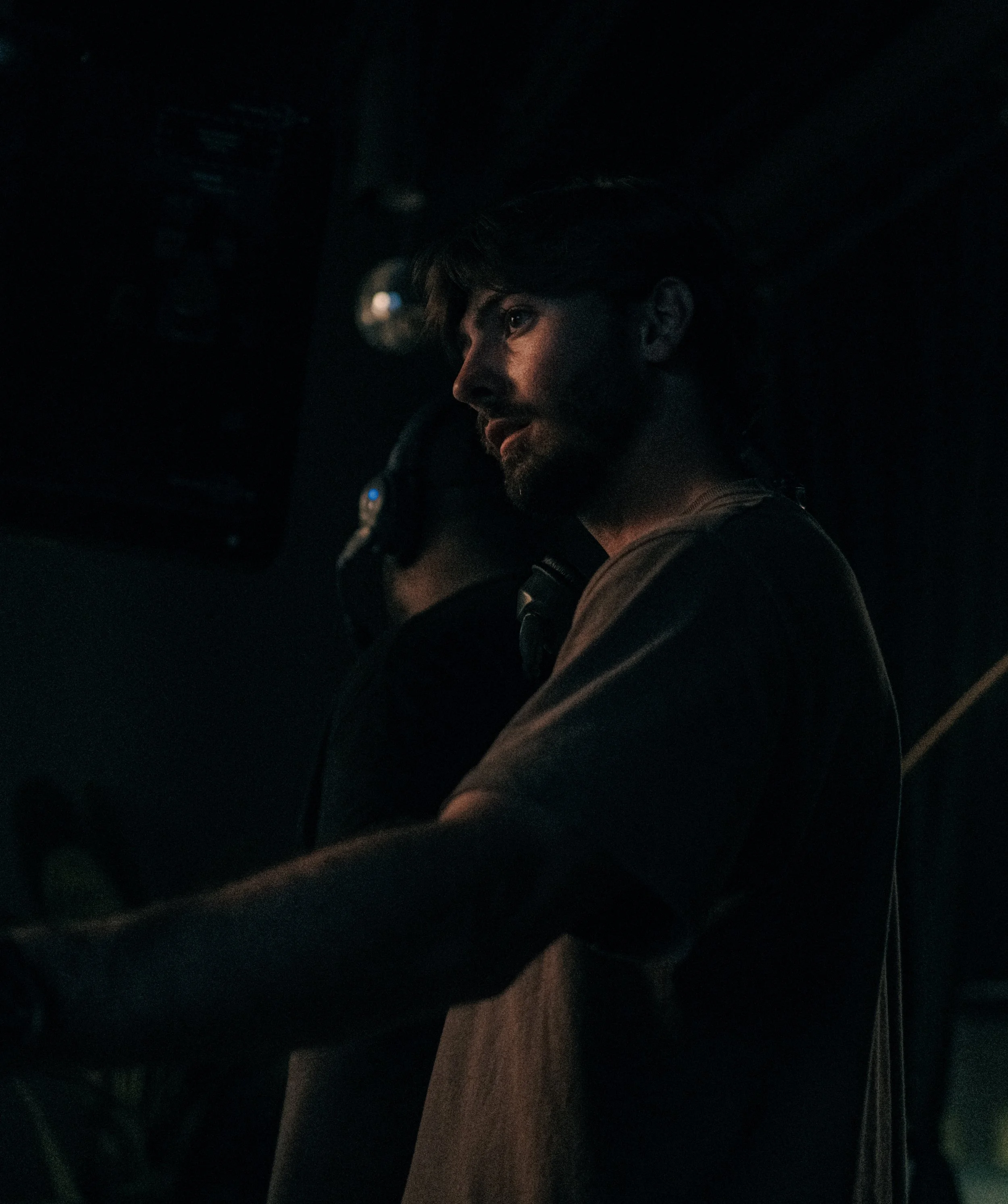 A man with a beard and dark hair, wearing a gray t-shirt, is looking to his right in a dark environment, possibly a music concert or club.