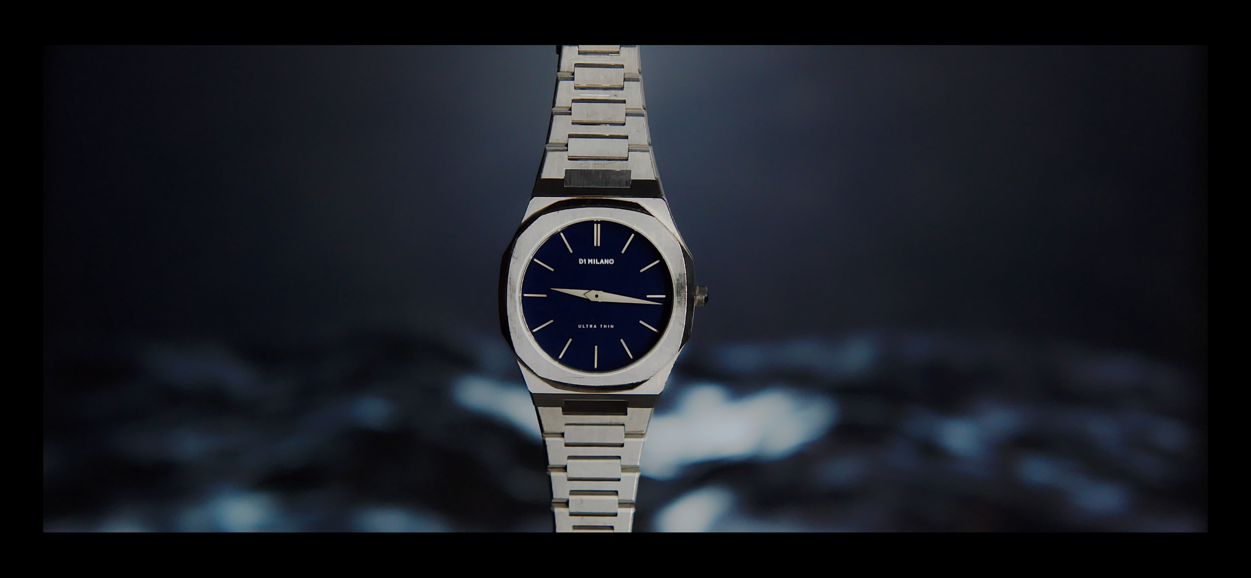 A silver wristwatch with a dark blue face, silver hour and minute hands, and a silver strap, floating above a dark, wavy background.