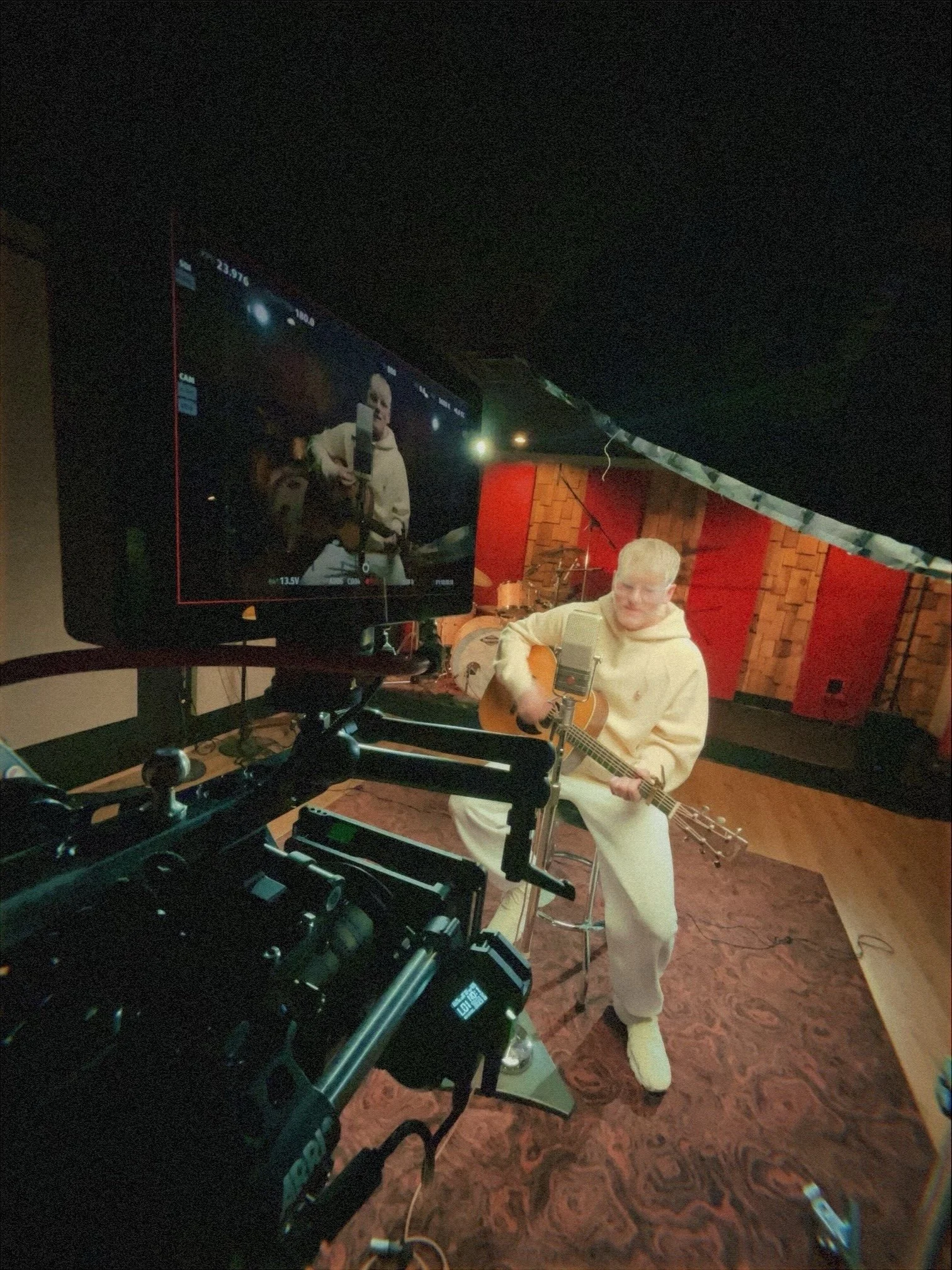 A person in a cream-colored hoodie and sweatpants is playing an acoustic guitar on a stage, with a microphone in front. A camera is recording the person, and the camera's monitor shows a live view of the person performing.