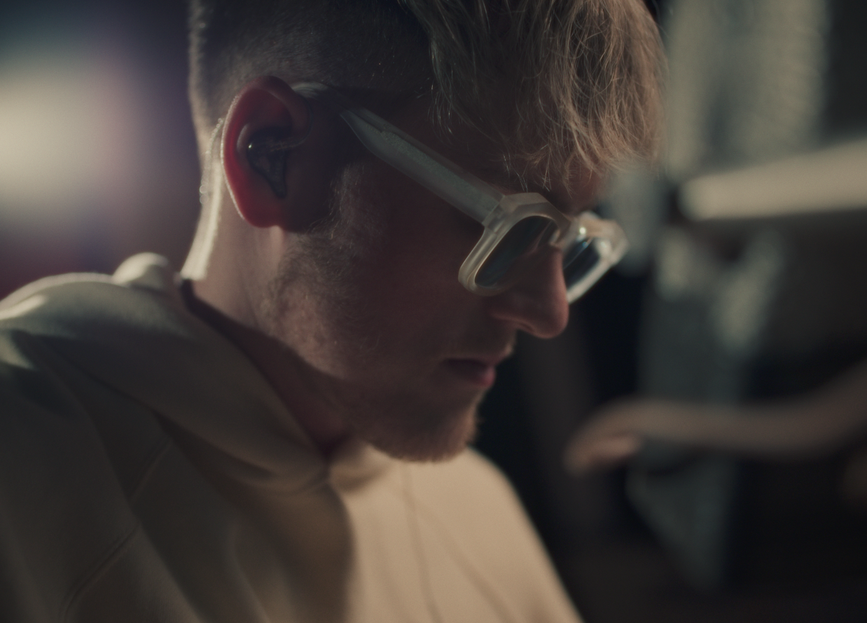 Close-up of a young man wearing glasses and a hoodie looking down, with earphones in his ears. The background is blurred.