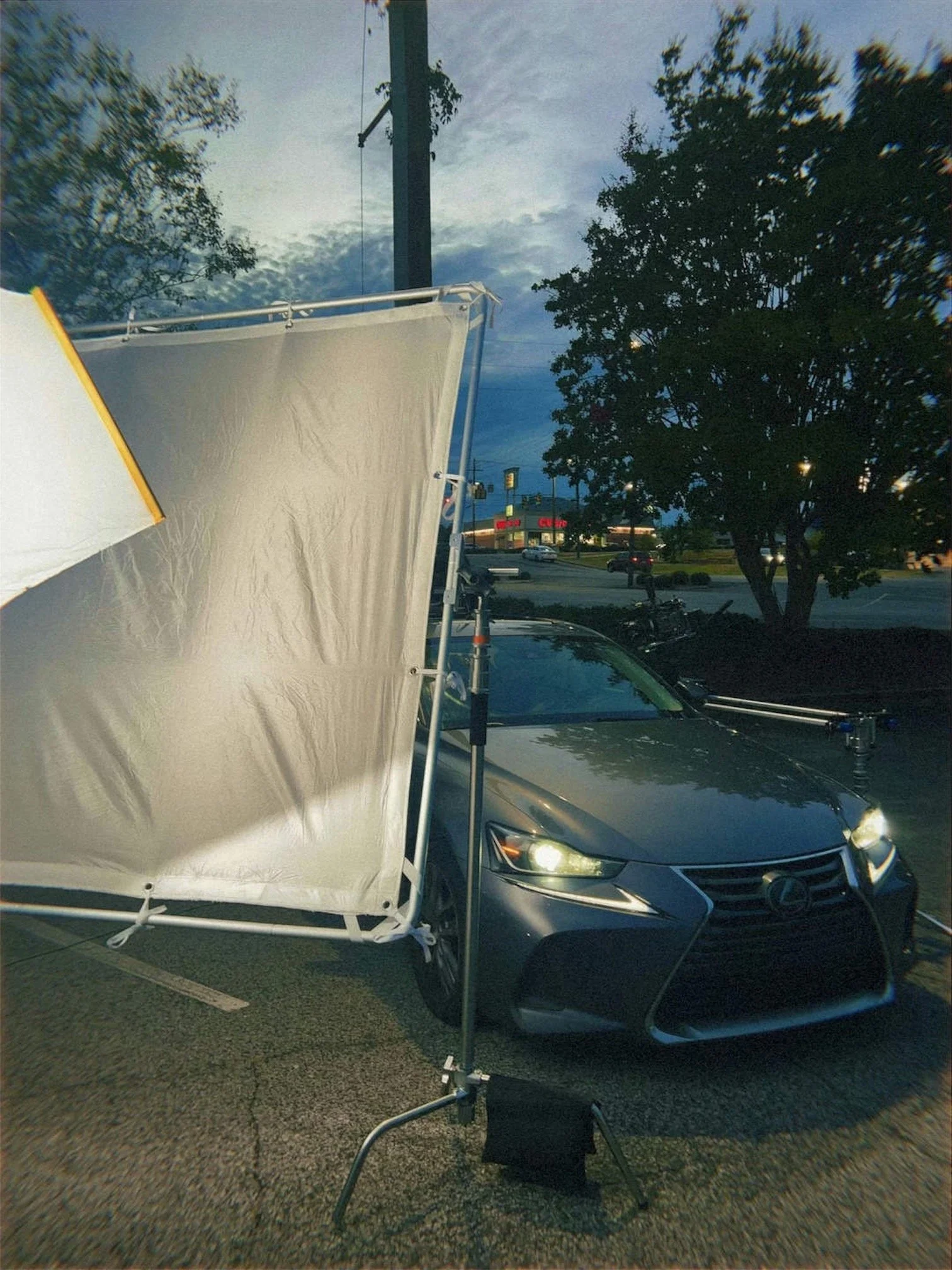 A nighttime scene in a parking lot showing a film or photography setup with a large white reflector or diffuser, a gray Lexus car with its headlights on, and a large tree in the background.