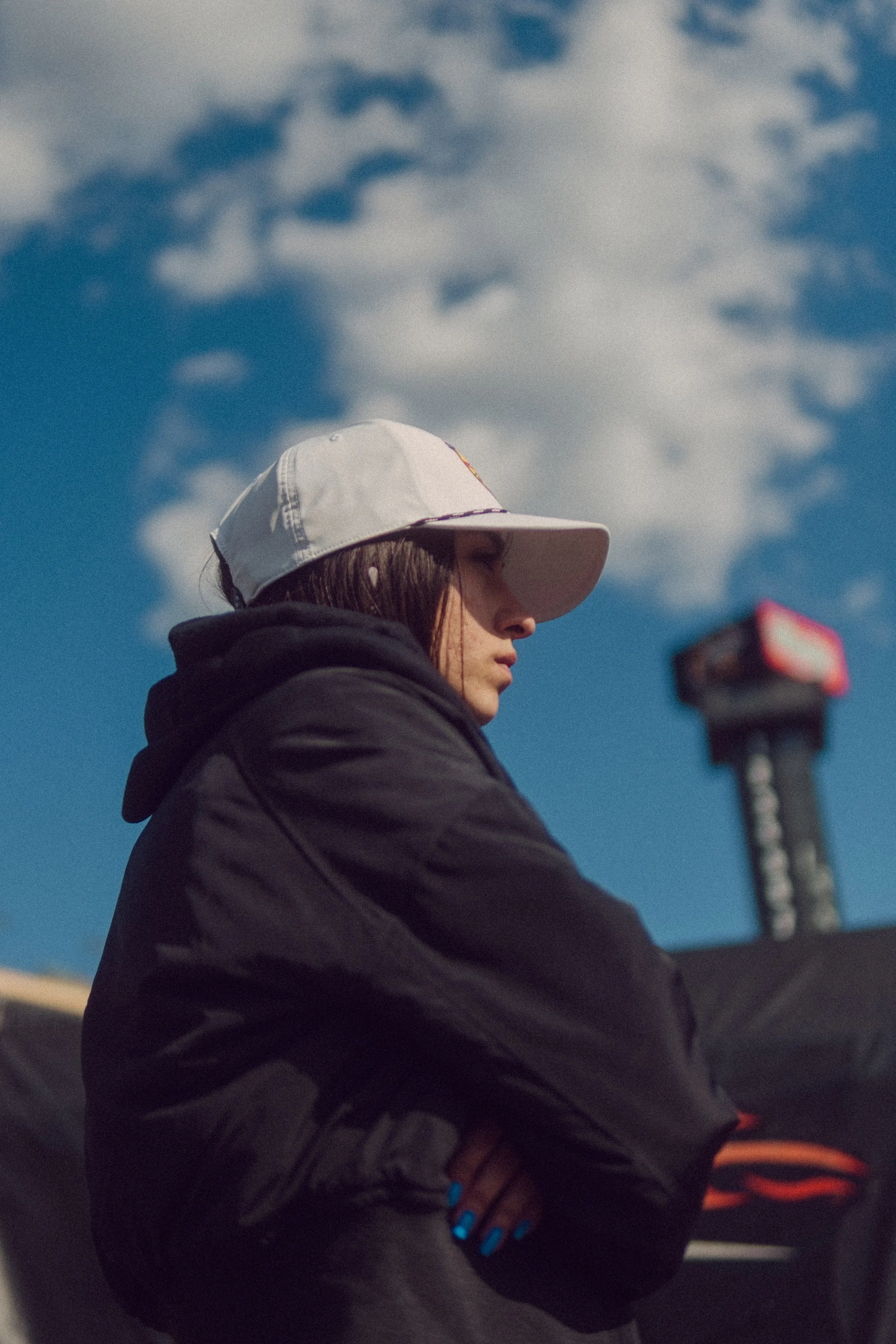A young woman with brown hair wearing a white cap and black hoodie, standing outdoors with crossed arms, under a partly cloudy blue sky.