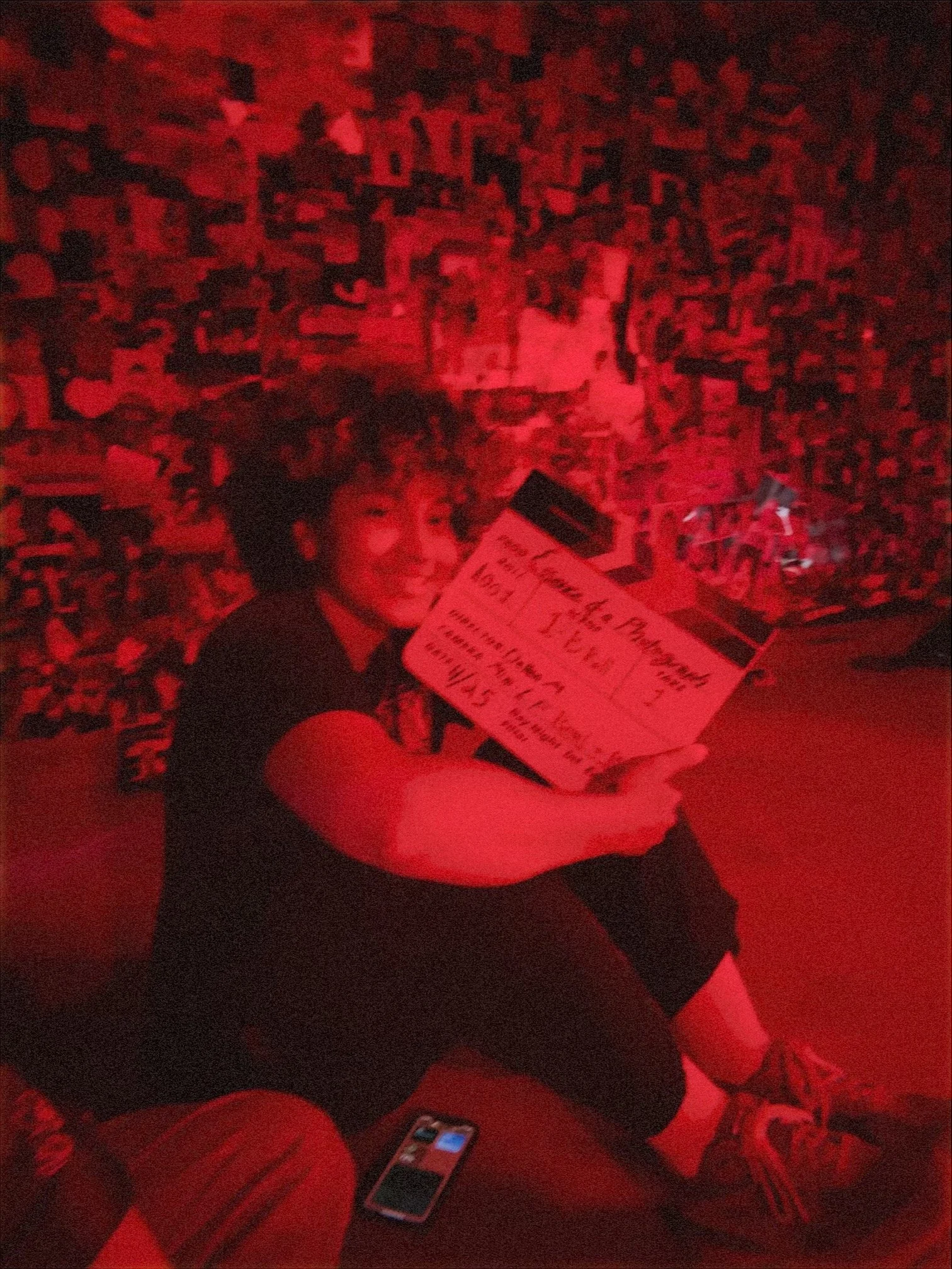 Person sitting in a dimly lit space with red lighting, holding a sign or poster with handwritten text, with a background covered in numerous posters or images.