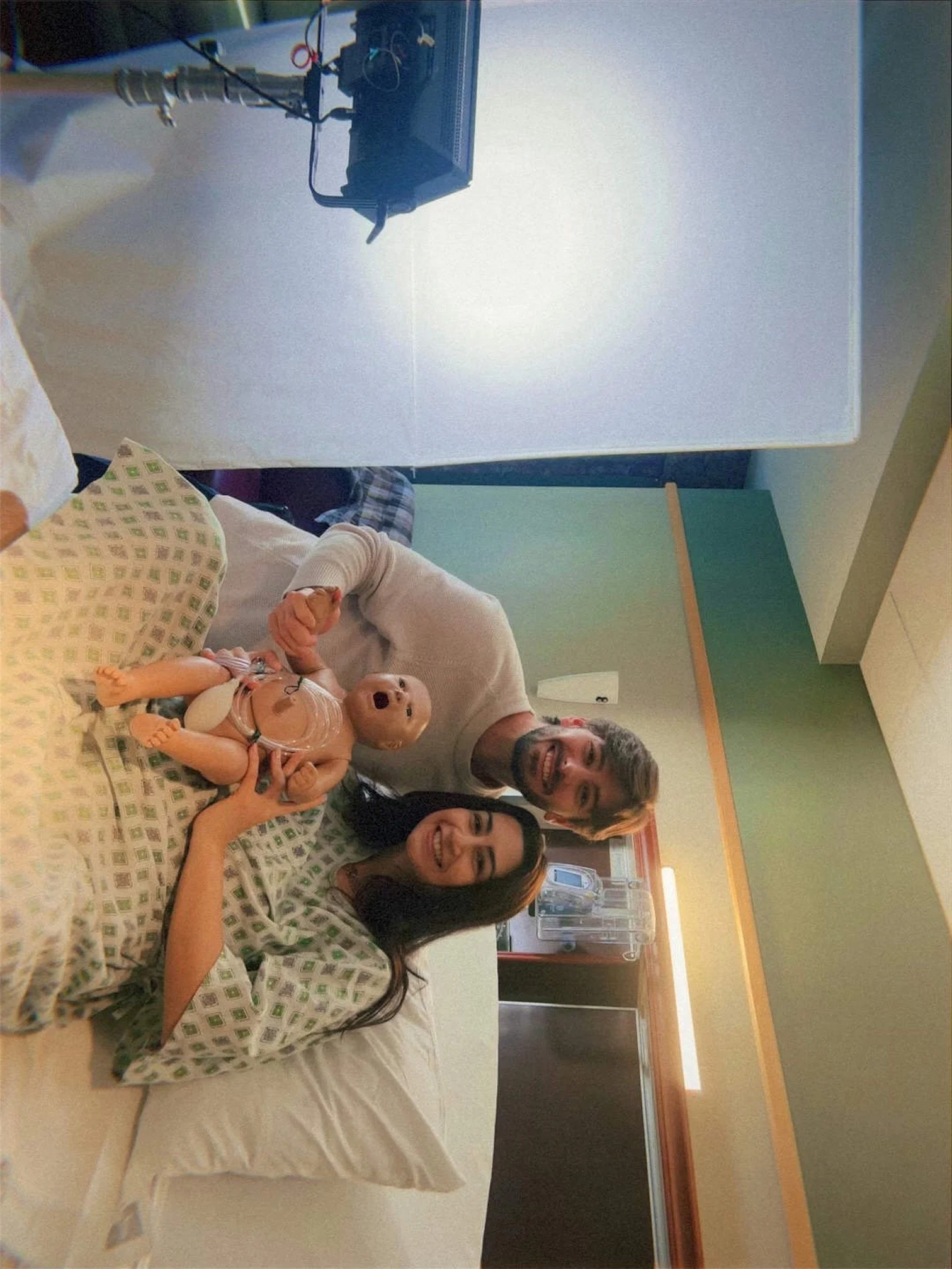 Two smiling hospital patients, a woman and a man, lying in a hospital bed holding hands with a doll and smiling at the camera.