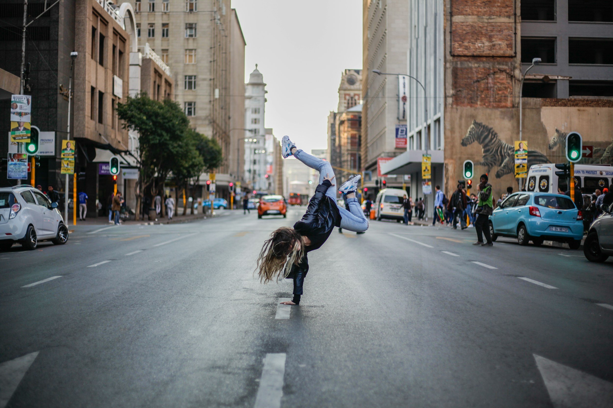 A woman in a black jacket and blue jeans is performing a handstand on a city street, with her legs bent backwards. The street is busy with cars and pedestrians, and there are buildings on either side, some with murals of zebras on the walls.