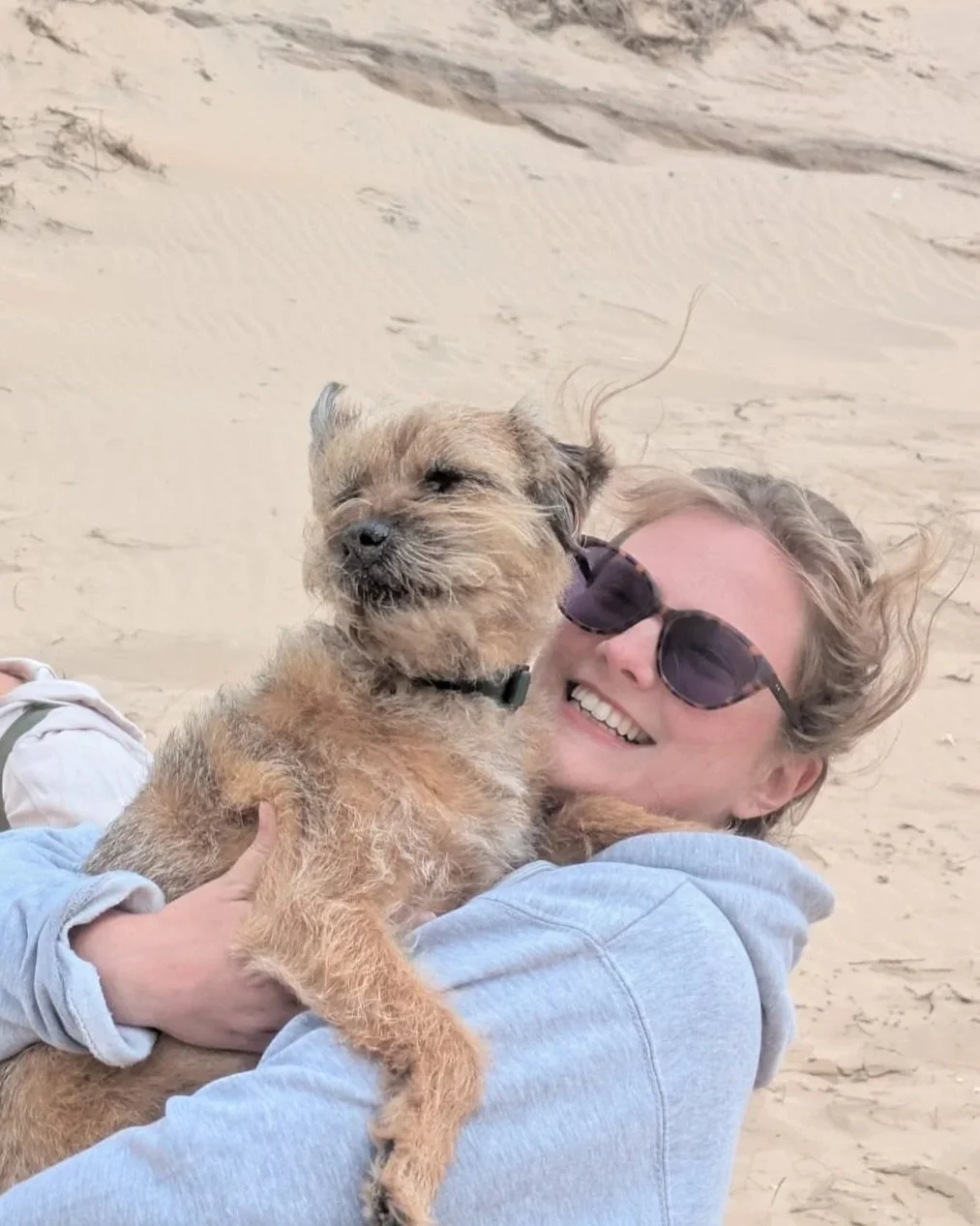 A woman with wavy blonde hair, sunglasses, and a gray hoodie smiling while holding a small scruffy dog, both on a sandy beach.