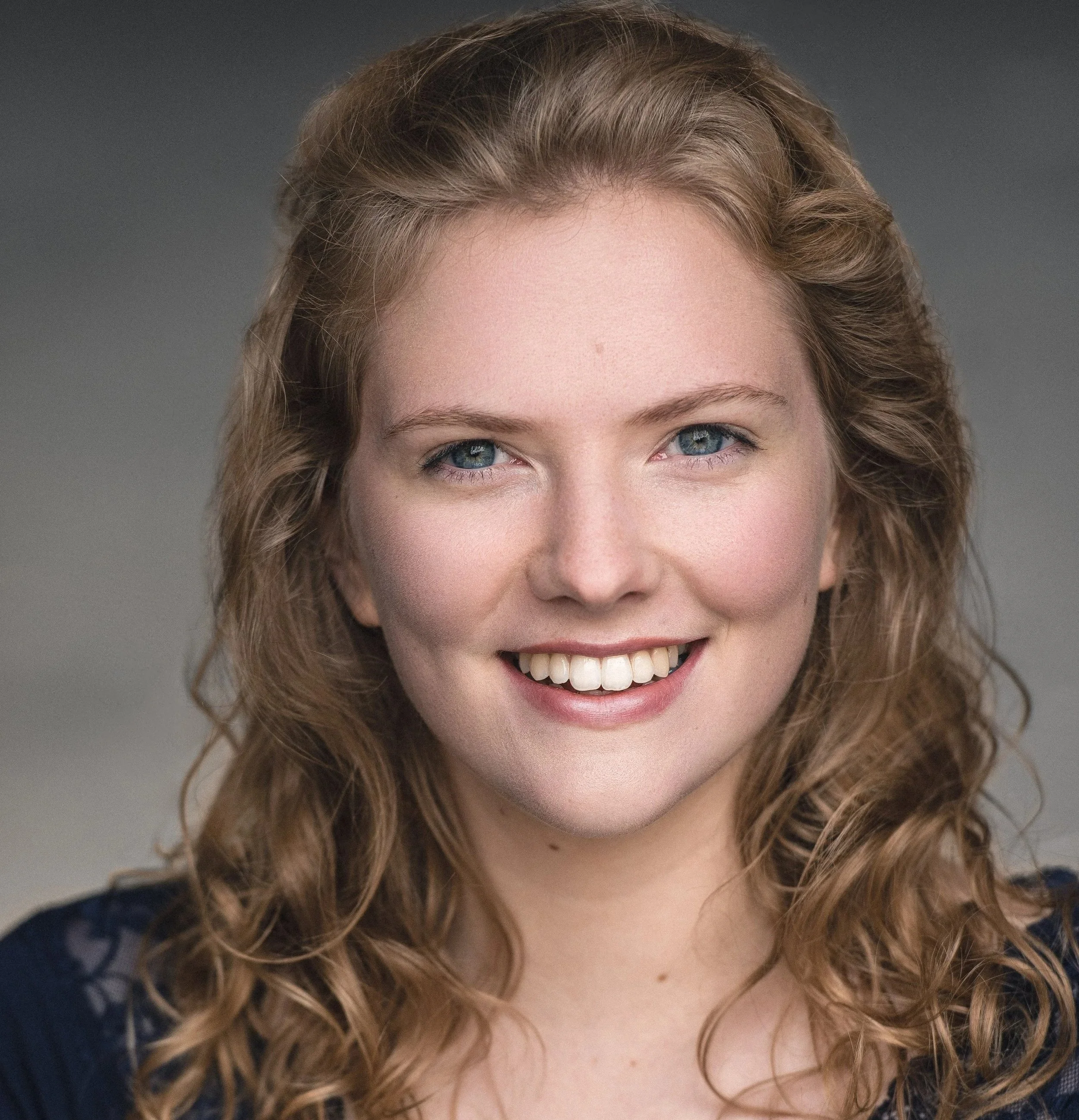 Close-up of a smiling woman with light brown, curly hair and blue eyes, wearing a dark top with lace detail on the shoulders.