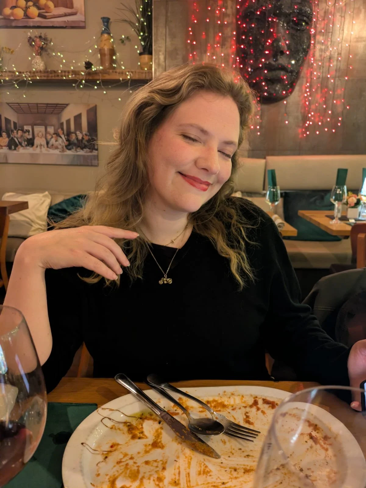 A woman with light brown wavy hair and a black top smiling with her eyes closed at a dining table, with a partly eaten plate of food in front of her and glasses of water in the background, in a restaurant decorated for Christmas with red string lights and a large decorative face on the wall.