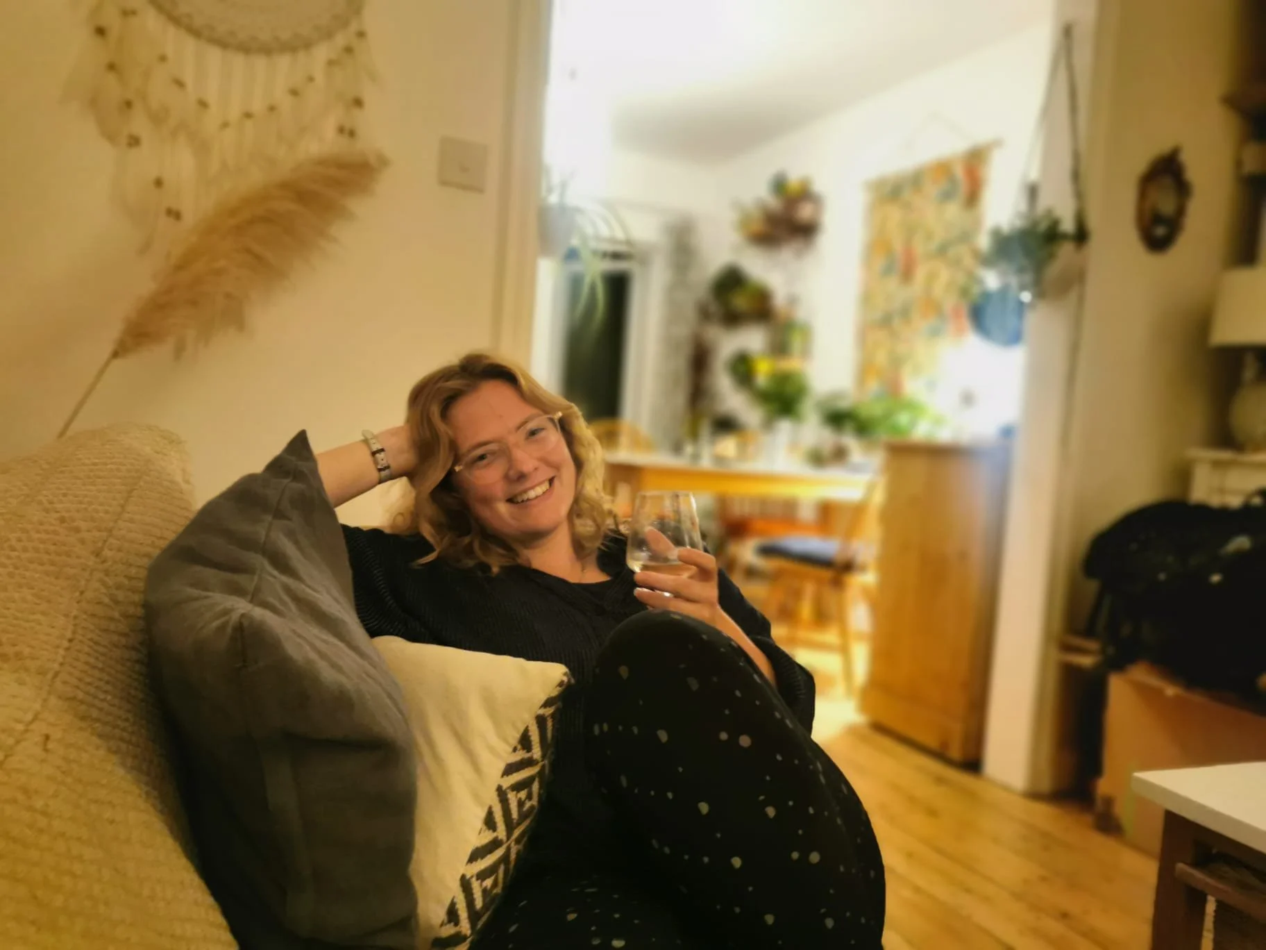 A woman with curly blonde hair and glasses is relaxing on a beige couch, smiling and holding a glass of wine in a cozy, well-lit living room with a kitchen in the background.