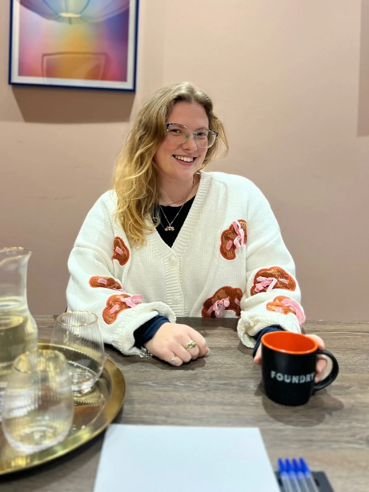 A woman with glasses and wavy blonde hair smiling while sitting at a table with a black mug that has 'FOUNDRY' written on it and a gold tray with glasses of water in front of her.