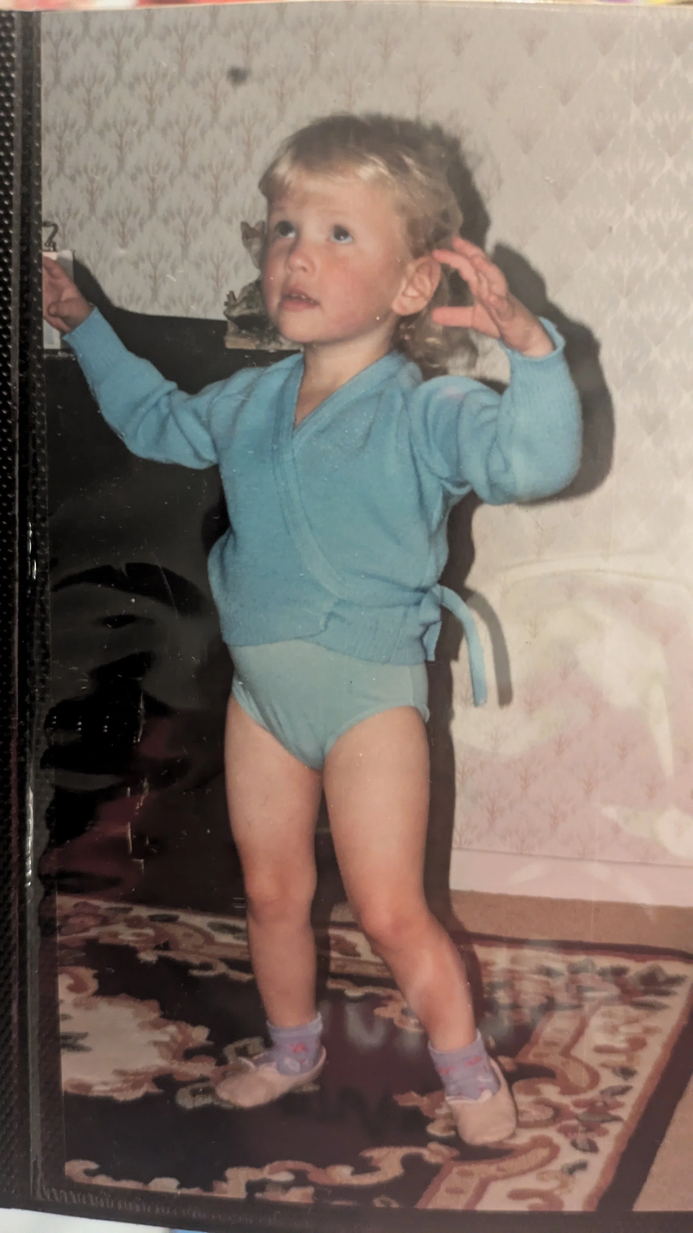 Child in a blue sweater, underwear, sneakers, and socks standing on a patterned rug, in a room with vintage wallpaper, holding onto a piece of furniture.