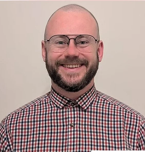 A man with glasses, a beard, and a shaved head smiling, wearing a checkered shirt, standing against a plain light-colored background.
