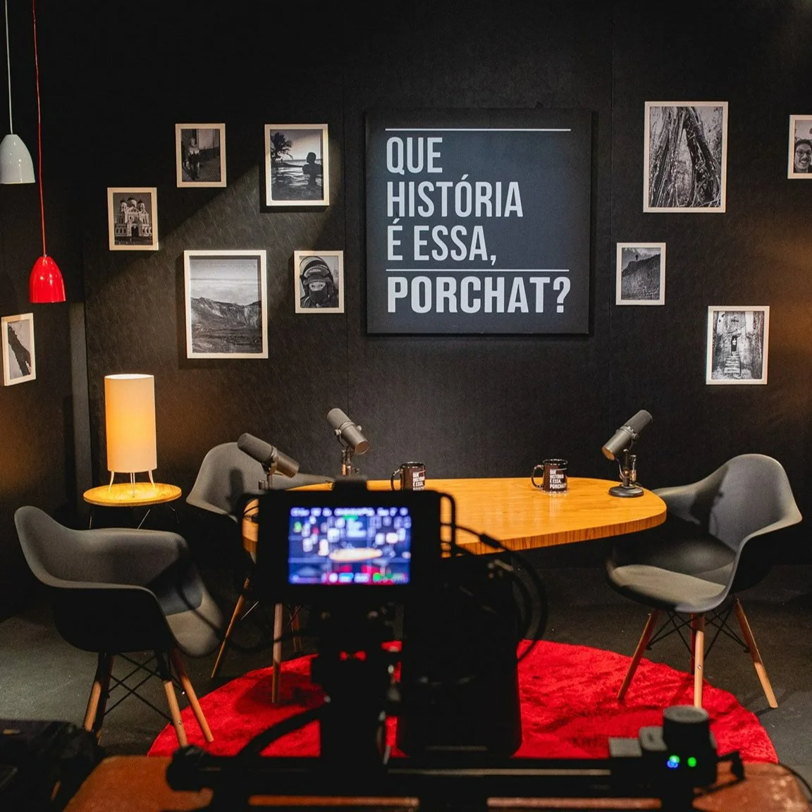 A recording studio setup with three gray chairs around a wooden table, black and white photos on a black wall, a large sign in Portuguese asking "What is this story?", a lamp, and microphones on the tables, with a camera in the foreground capturing the scene.