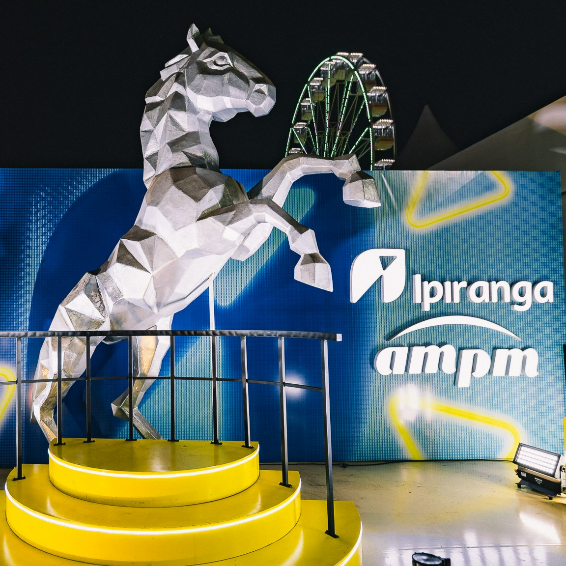 A large, geometric, silver unicorn statue on a yellow circular platform with a black railing. Behind it, a blue backdrop displays the logos 'Ipiranga' and 'ampm,' with a lit-up Ferris wheel in the background at night.