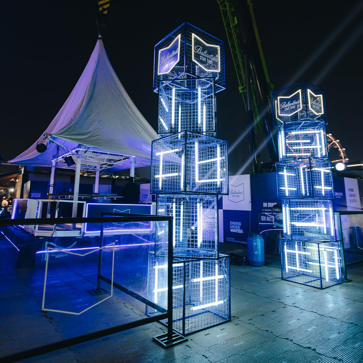 Night scene at a carnival or fair with illuminated blue and white neon cube structures and a white tent in the background.