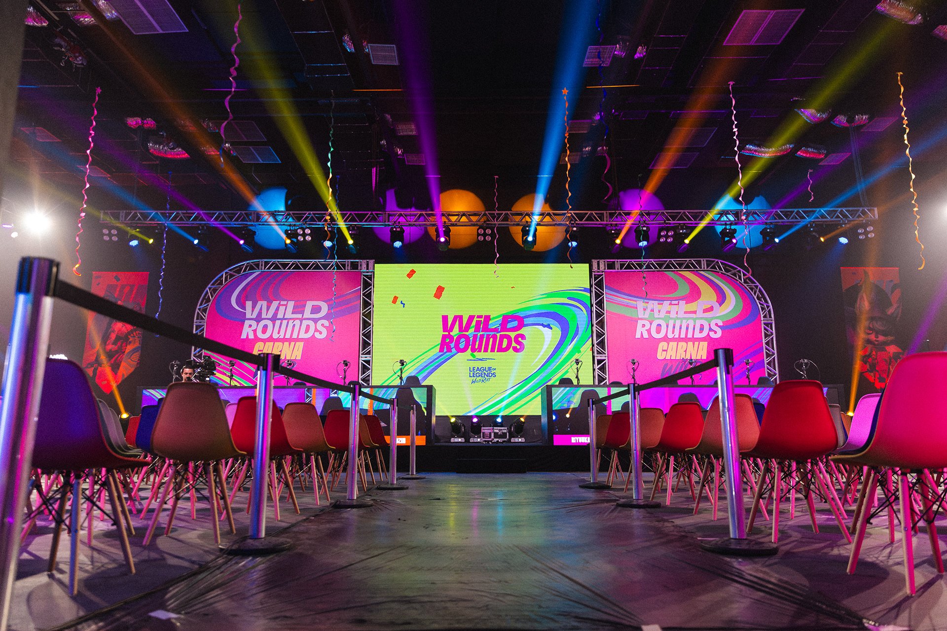 Palco iluminado com telões exibindo 'Wild Rounds', cadeiras dispostas em fileiras na frente do palco, iluminação colorida e confetes no ar, indicando preparação para evento de e-sports ou gaming.