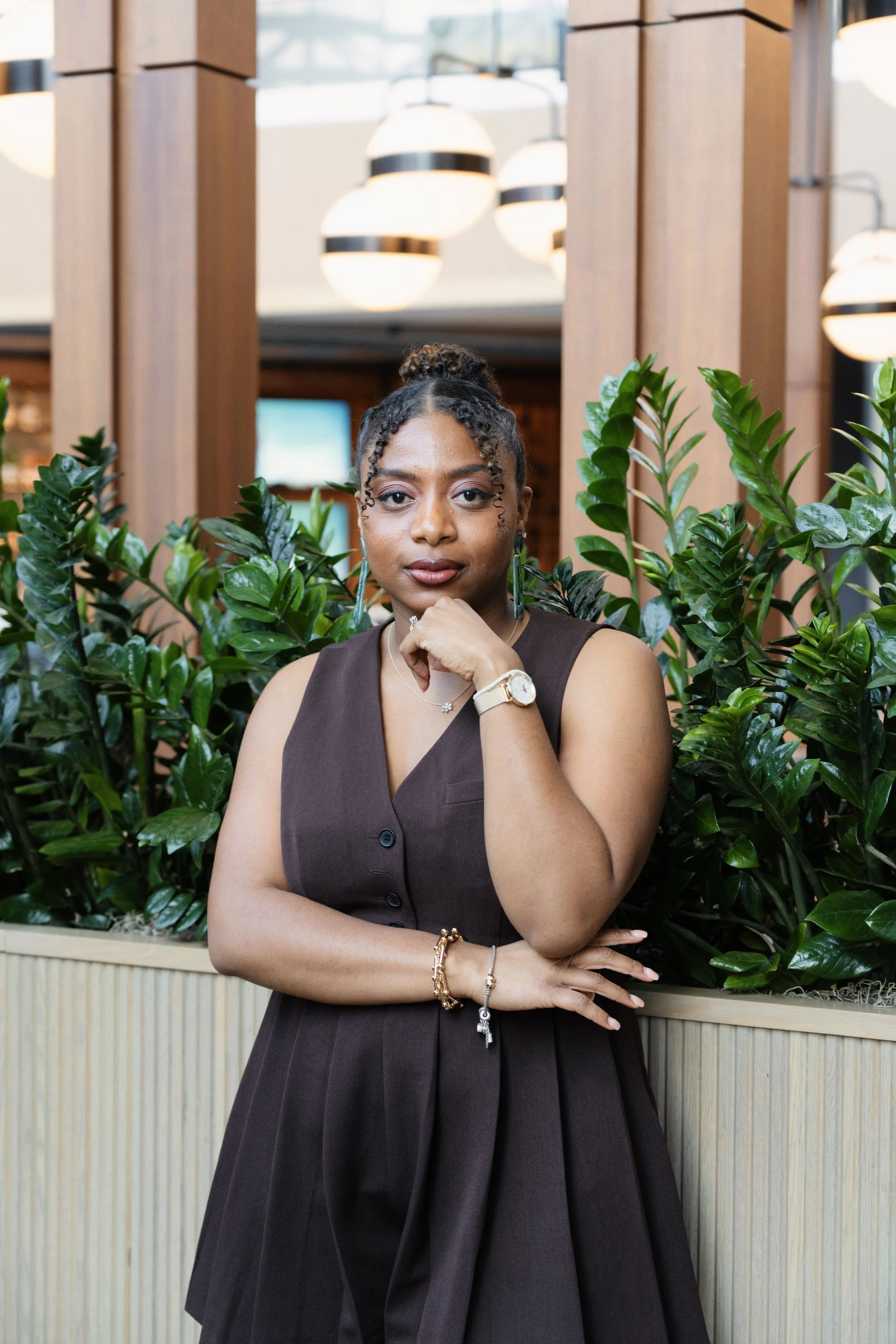 A woman wearing a sleeveless brown dress, standing in front of green plants and wooden decor, chin resting on her hand, posing confidently inside a modern indoor space.