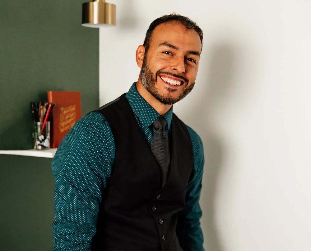 A smiling man with a beard and short hair, wearing a teal dotted shirt, dark vest, and tie, standing in a room with a green and white wall background.