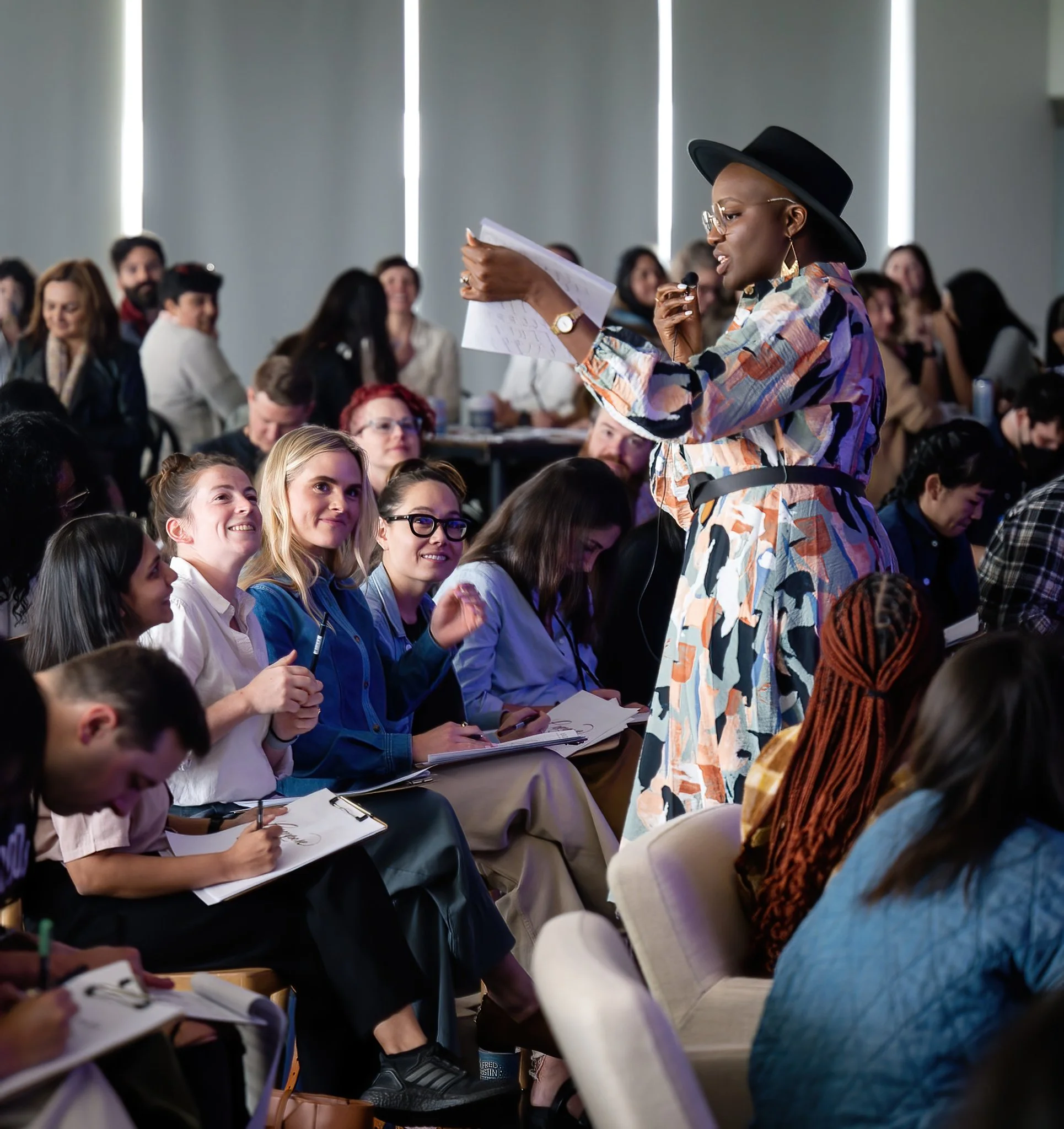 A woman in a colorful dress and black hat speaking to an audience during a conference or seminar.