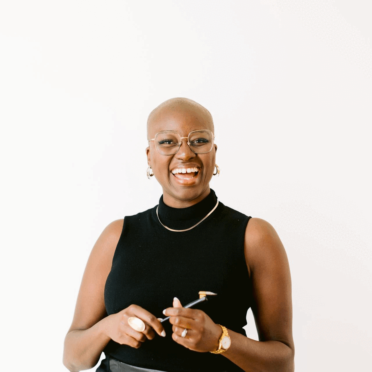 A woman with wearing glasses, and hoop earrings, wearing a black sleeveless turtleneck, smiling and holding a pen, standing against a white background.