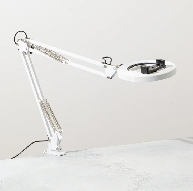 A white adjustable desk lamp with a magnifying glass, positioned on a white surface against a plain light gray background.