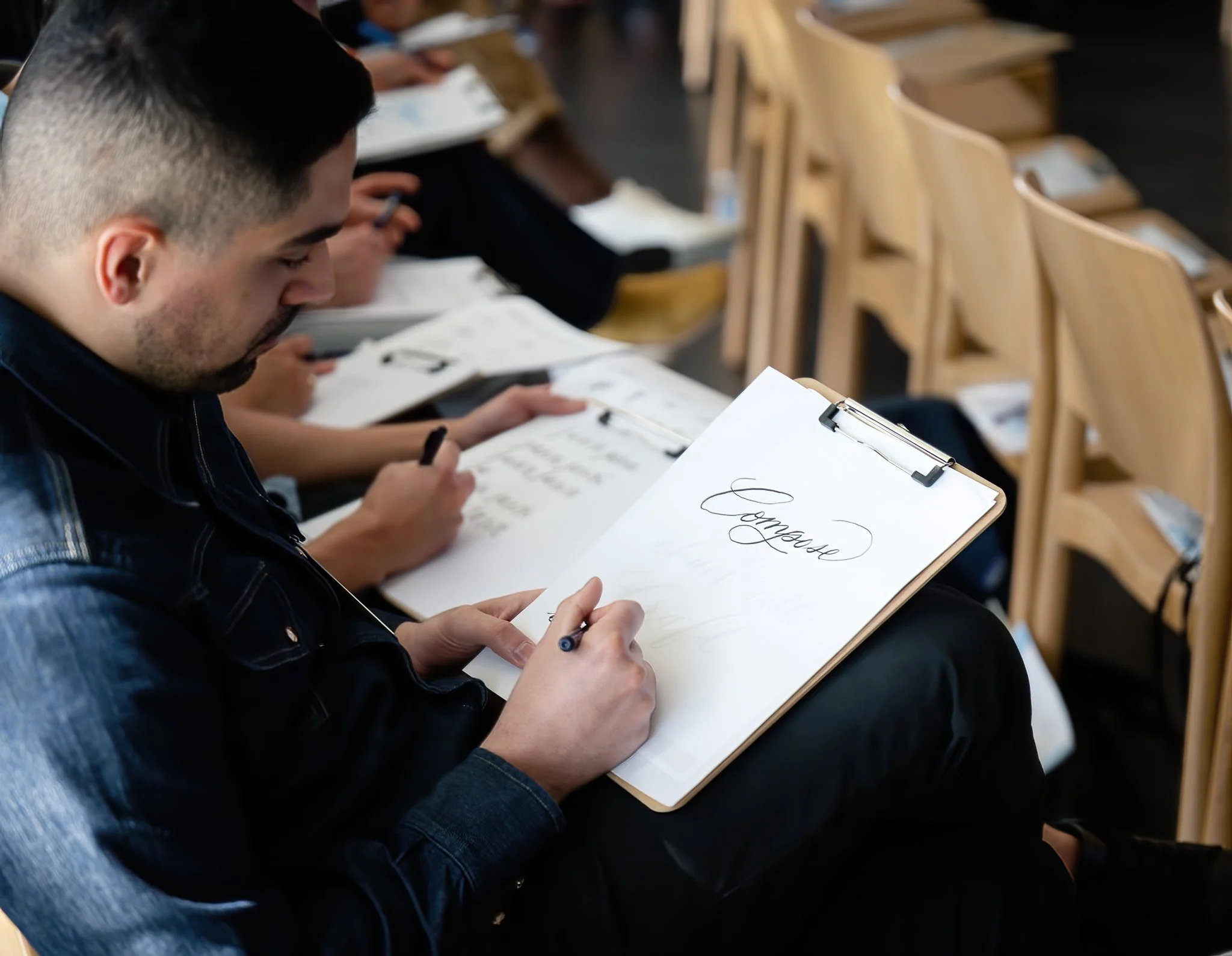 Man taking notes on a clipboard with the word 'Envelope' in calligraphy, seated in a classroom with other students writing and reading.