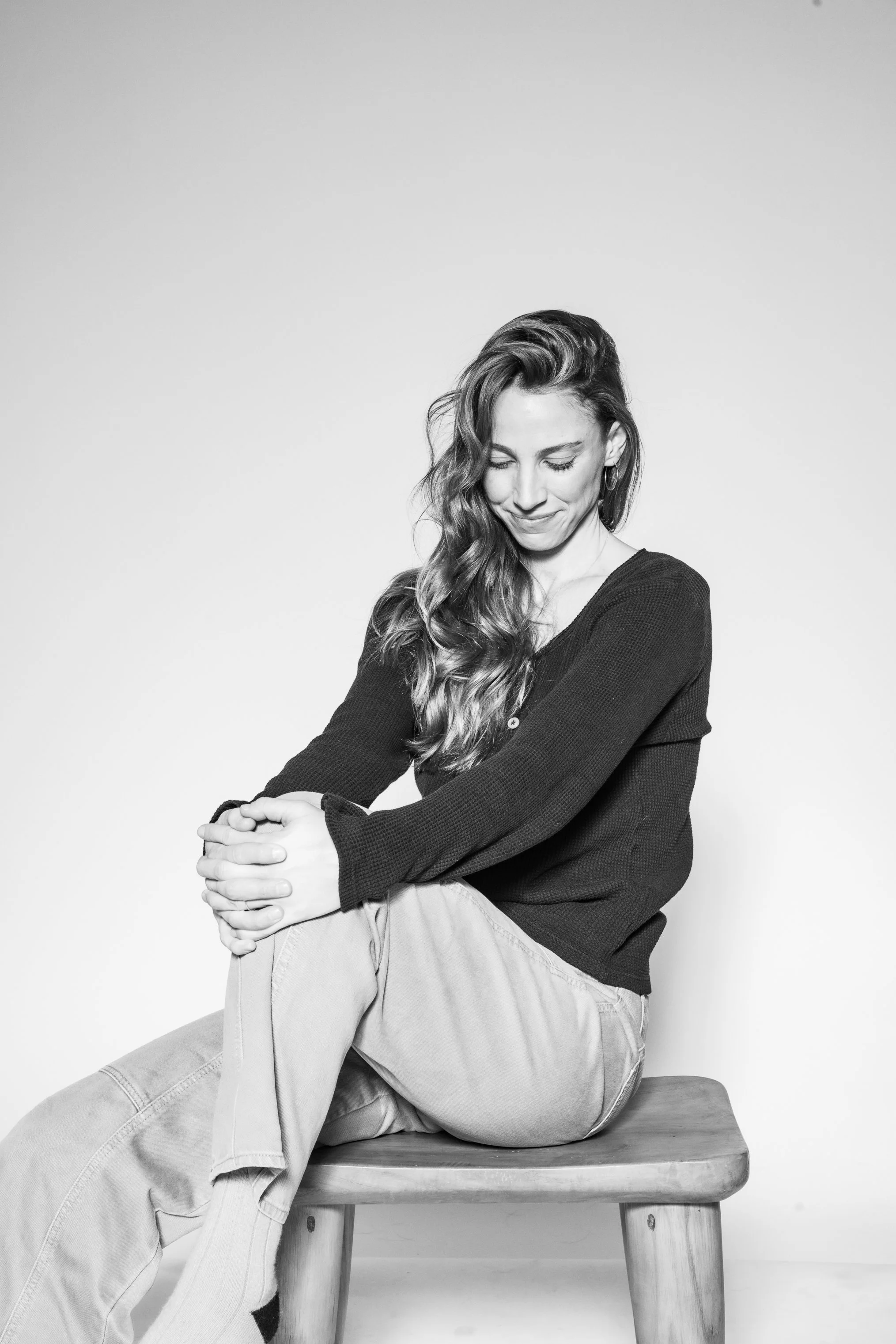 A woman with long wavy hair sitting on a wooden stool, smiling softly, wearing a dark long-sleeve shirt and light-colored pants, against a plain white background.