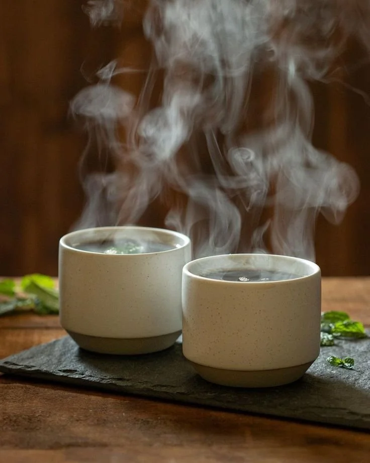 Two steaming cups of hot tea or coffee on a dark slate tray, with a wooden background.