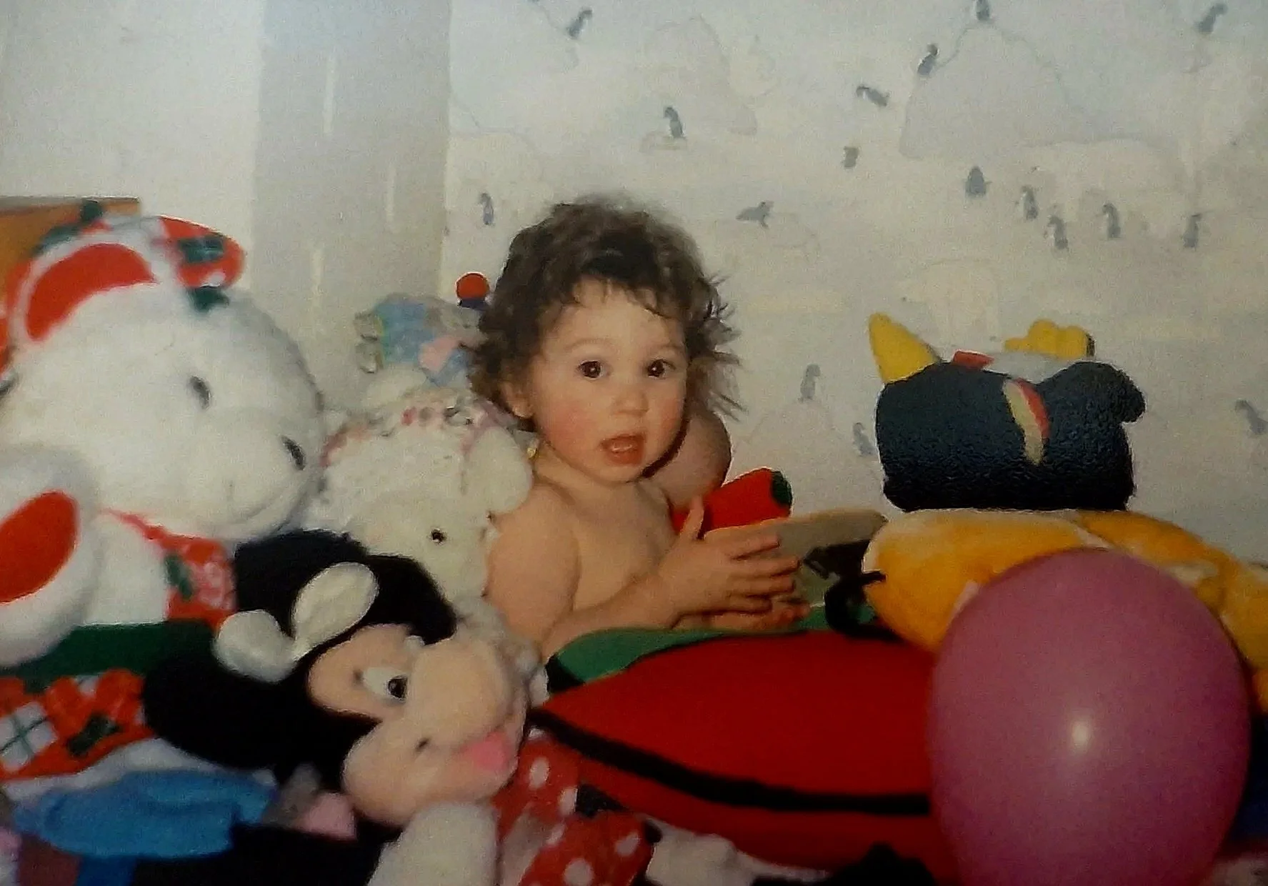 Me as a small child sitting up in bed, surrounded by stuffed toys and a balloon for some reason. Holding a book and looking autistictally at the camera.