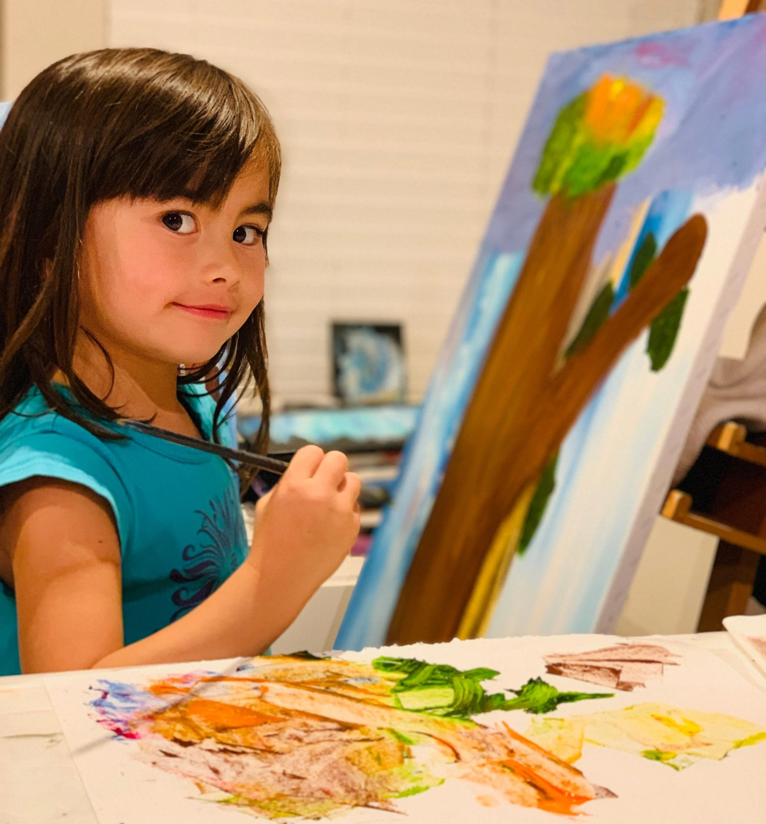 Young girl with brown hair wearing a blue shirt painting on a canvas with a brush, smiling at the camera. Art Classes and Parties in Cloverdale, Surrey, Langley, White Rock, Richmond, and Vancouver