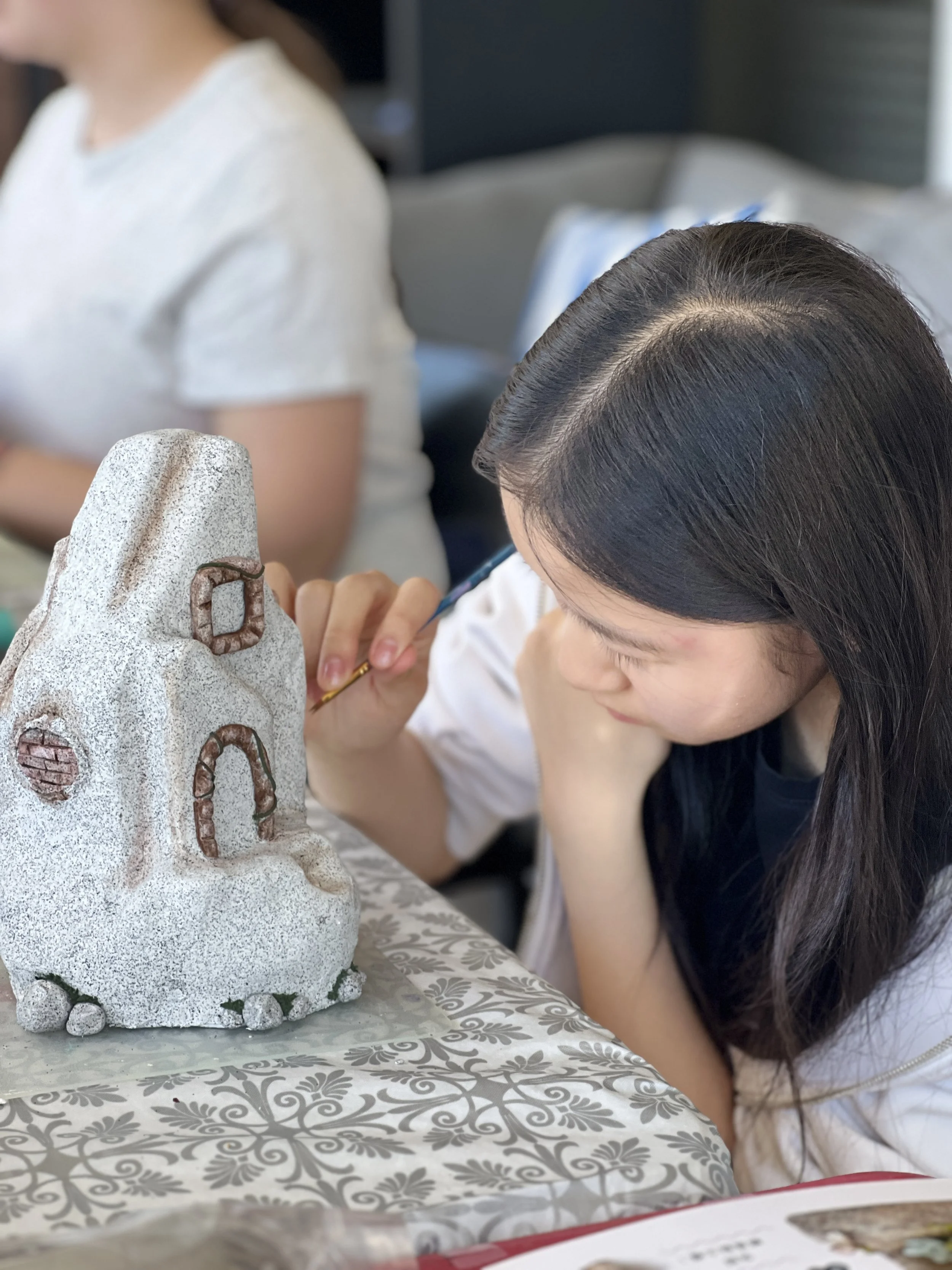 A woman is painting a stone sculpture resembling a face with windows and a door, sitting at a decorated table.  Art Classes, Workshops and Parties with Party with Laura. Available in Cloverdale, Bc Surrey, Langley, White Rock, Richmond, Vancouver, an