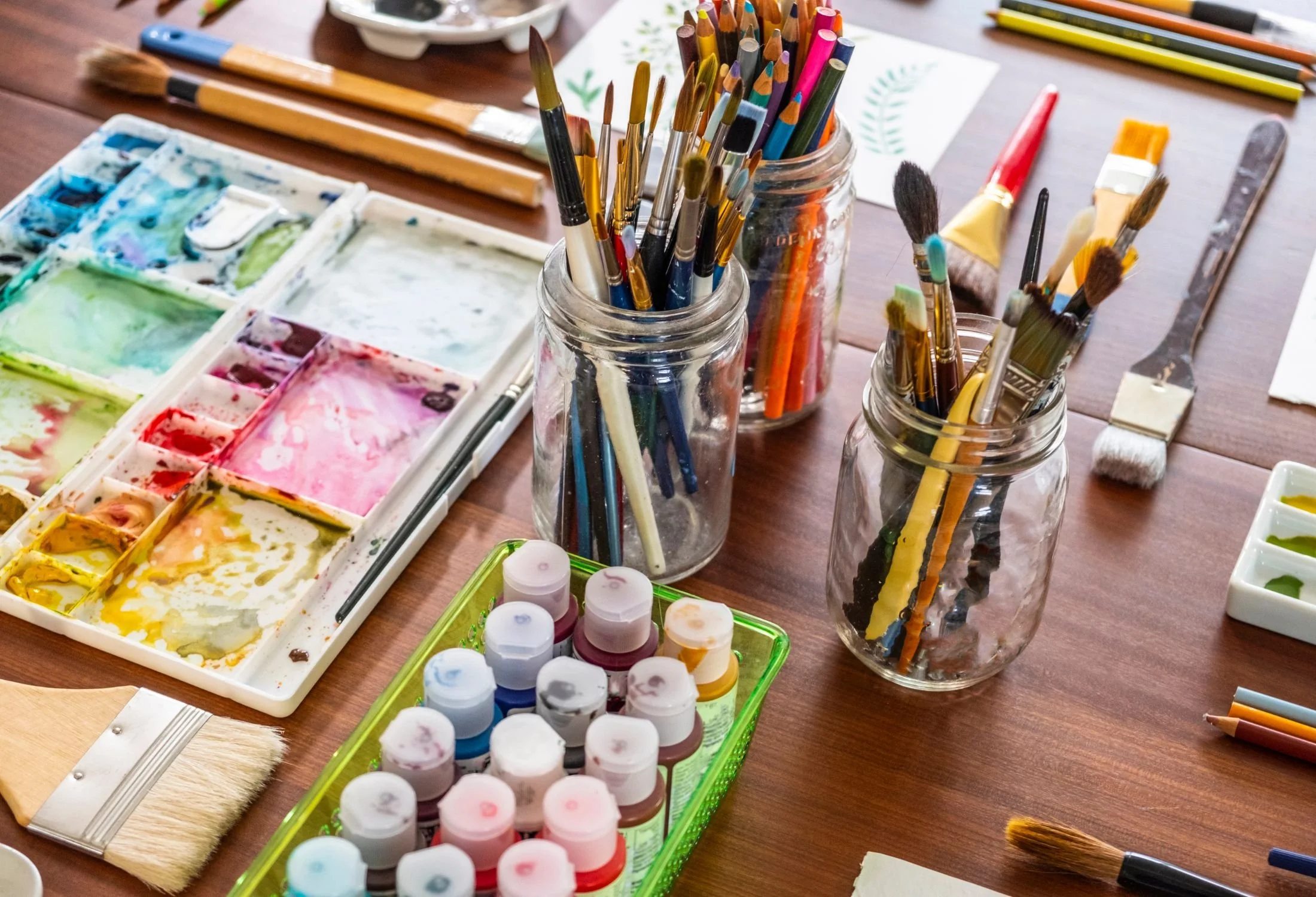 Art supplies including paint bottles, watercolor palette, paintbrushes in jars, colored pencils, and paper on a wooden table.  Art Classes, Workshops and Parties with Party with Laura. Available in Cloverdale, Bc Surrey, Langley, White Rock, Richmond