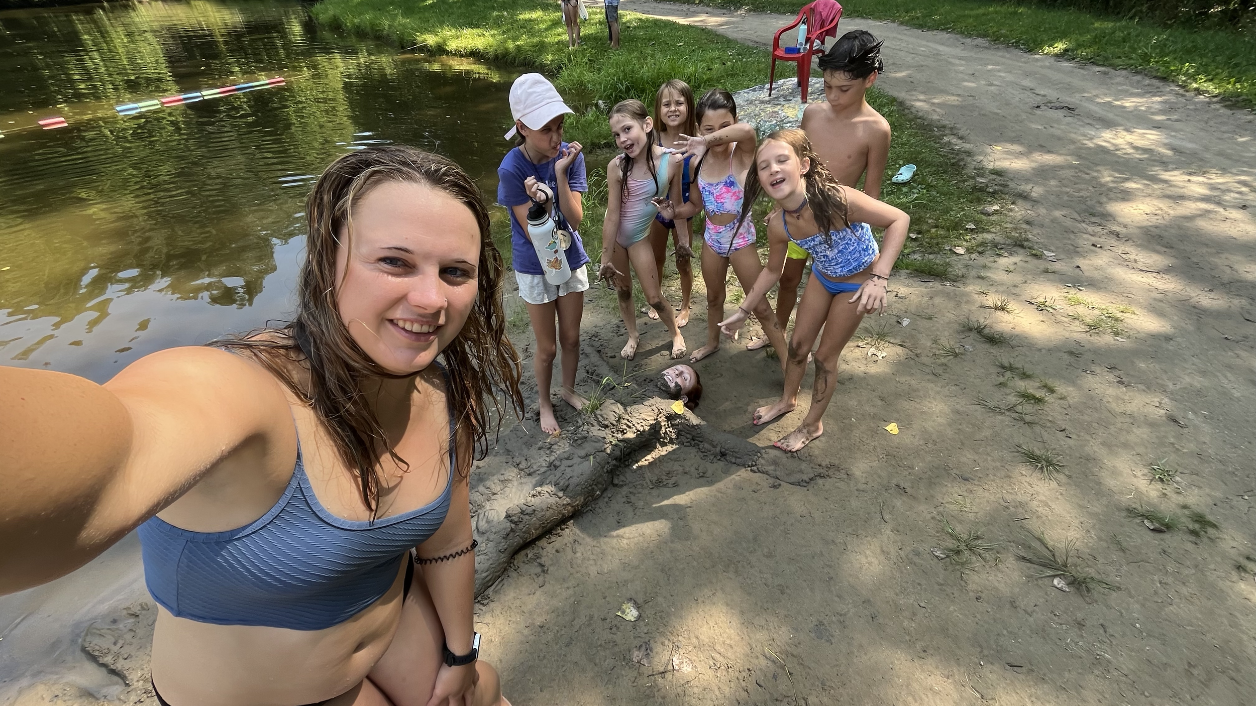 A group of children and a young woman smiling at the camera in a sandy outdoor area near a body of water, with some children playing and a person submerged in the sand.