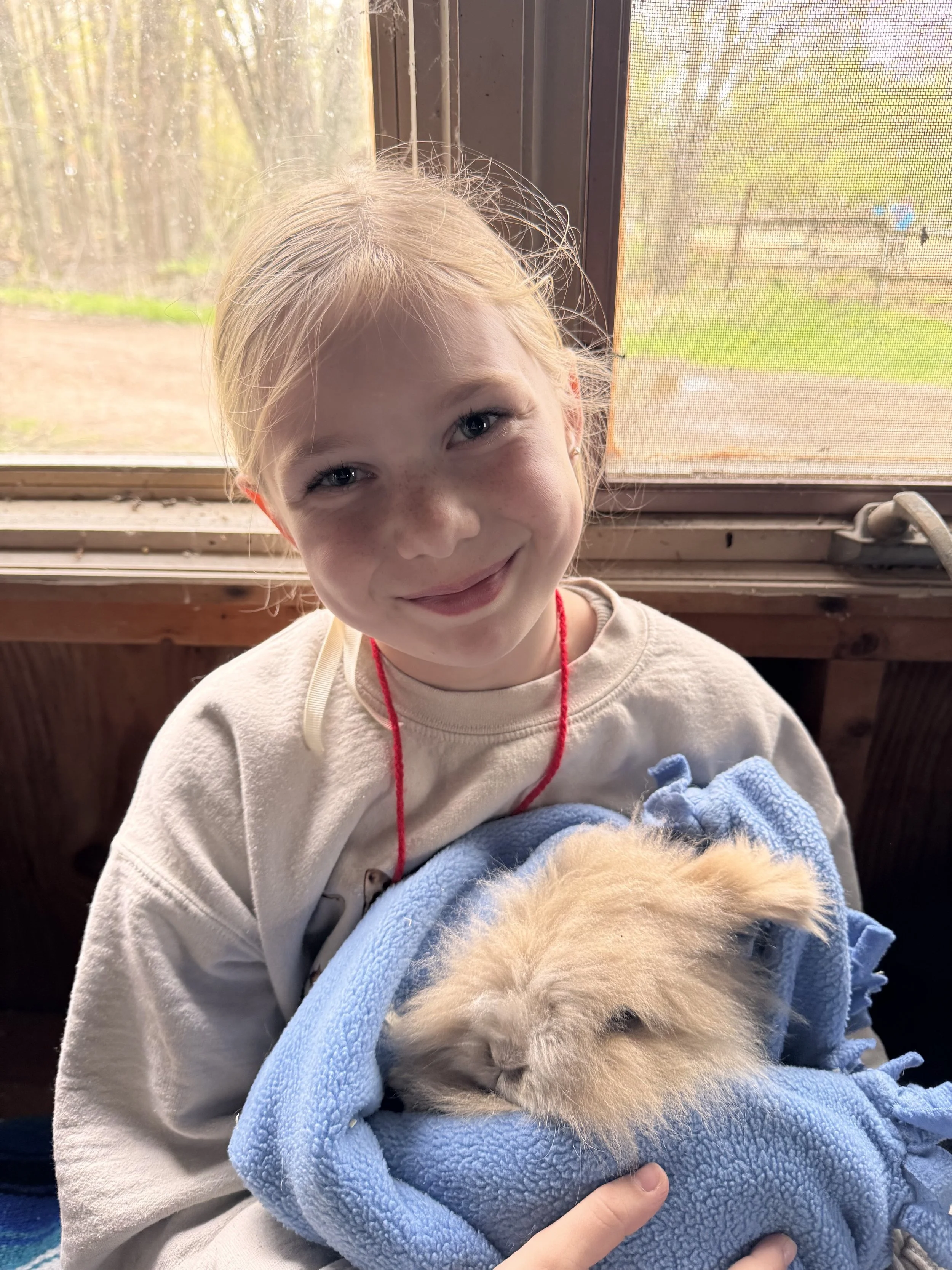 A young girl with red hair and a red necklace smiling while holding a small, fluffy, light-colored puppy wrapped in a blue blanket.