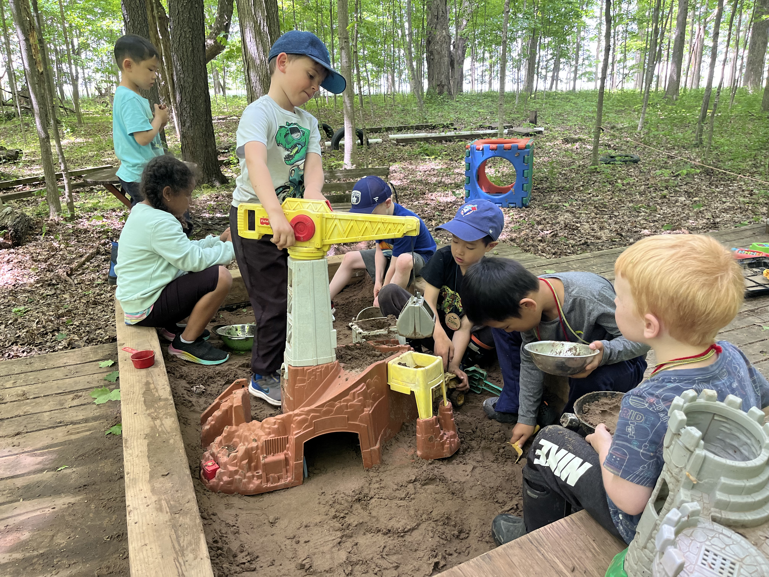 Children playing in a sandbox with toy construction equipment in a wooded outdoor area.