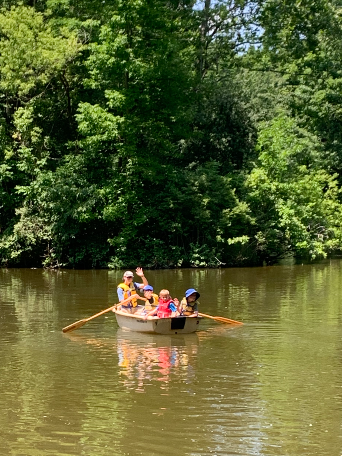A group of four children and one adult are in a small boat on a river, surrounded by green trees. The adult and one child are waving, and everyone is wearing life jackets.