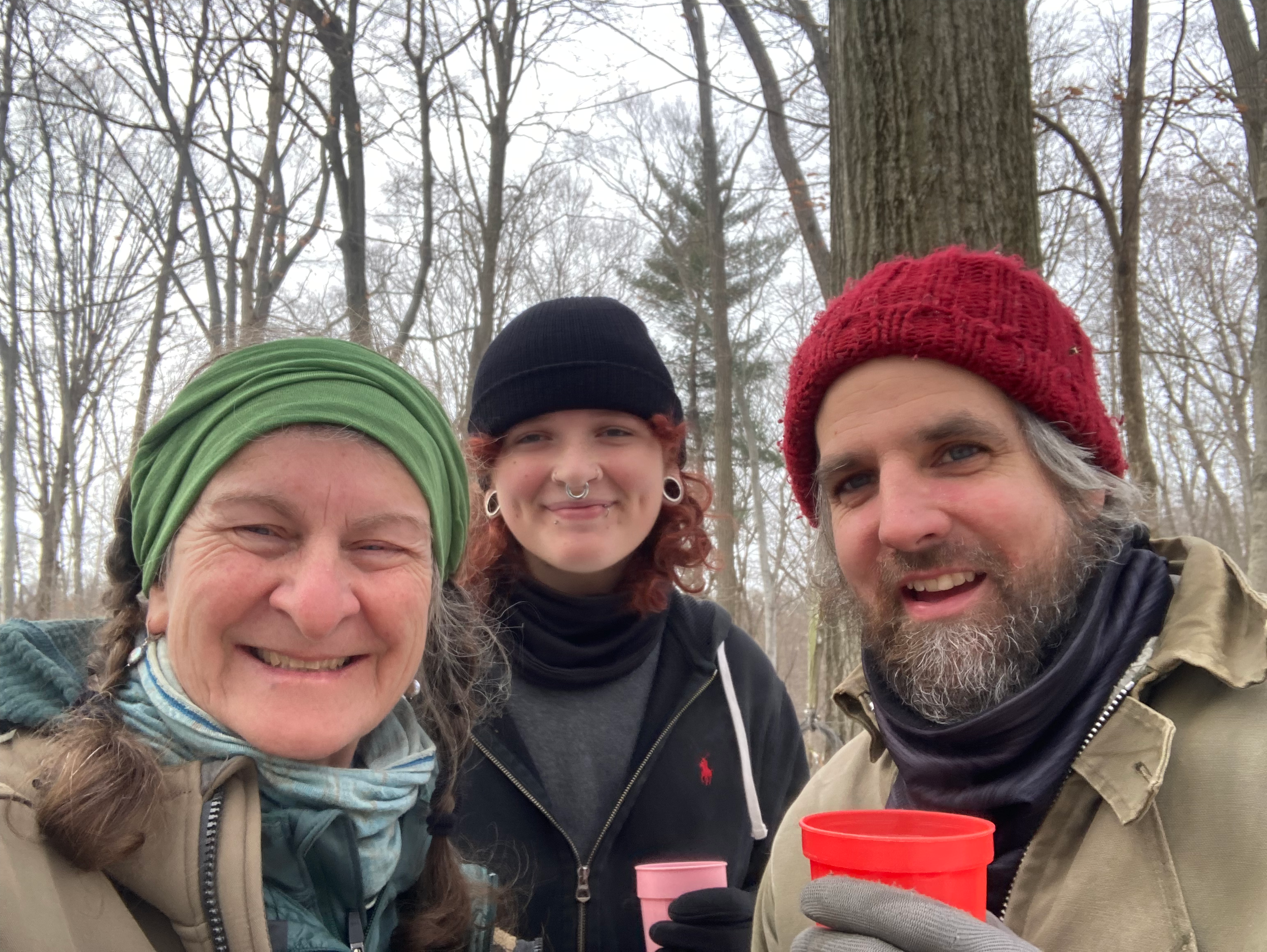 Four people smiling in a winter forest, wearing warm clothing and hats, with leafless trees and overcast sky in the background.