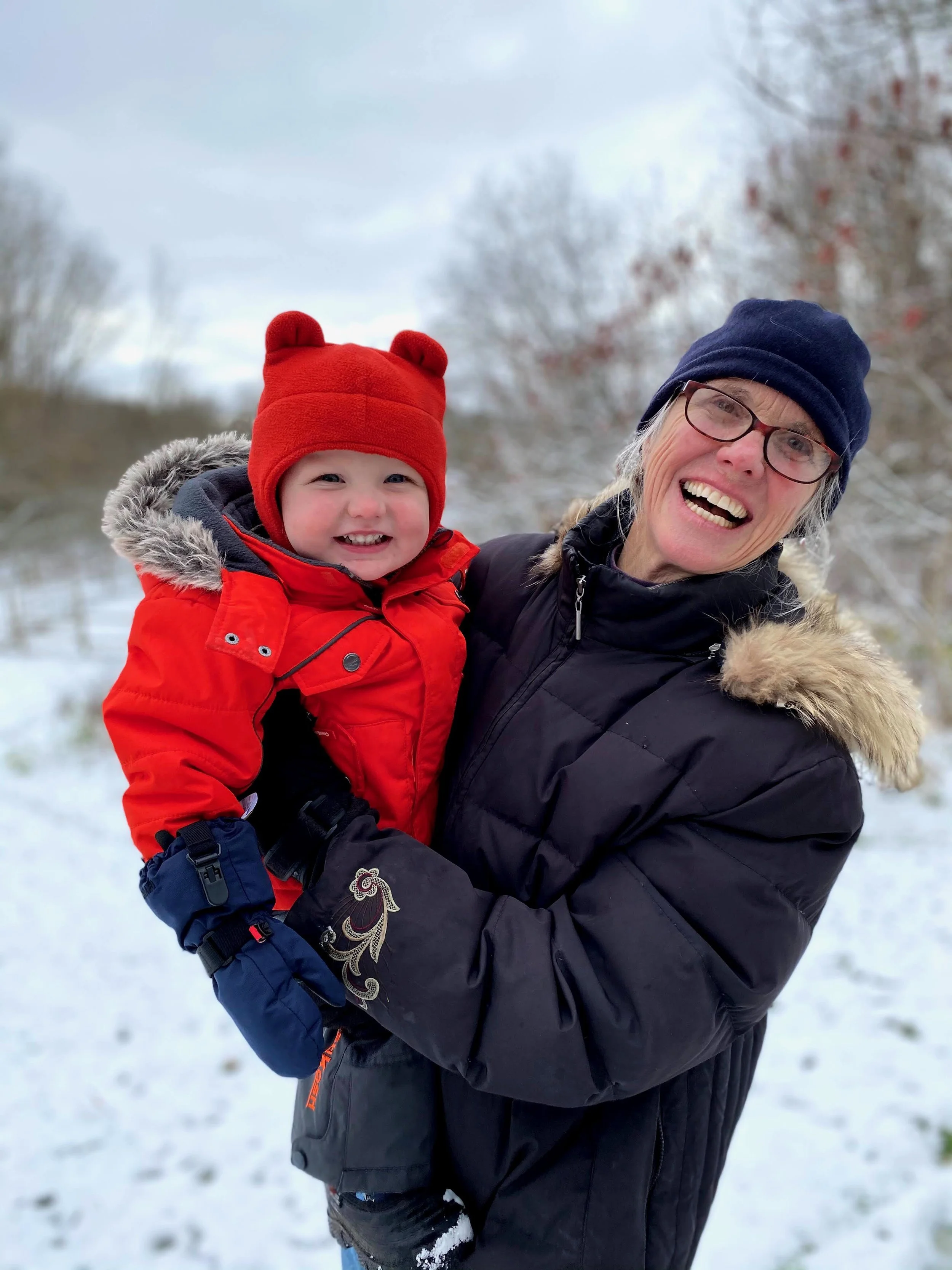 An elderly woman wearing glasses, a navy beanie, and a black winter coat with a fur-lined hood holding a smiling young child dressed in a red winter jacket, red hat with bear ears, and blue gloves outdoors in a snowy landscape.