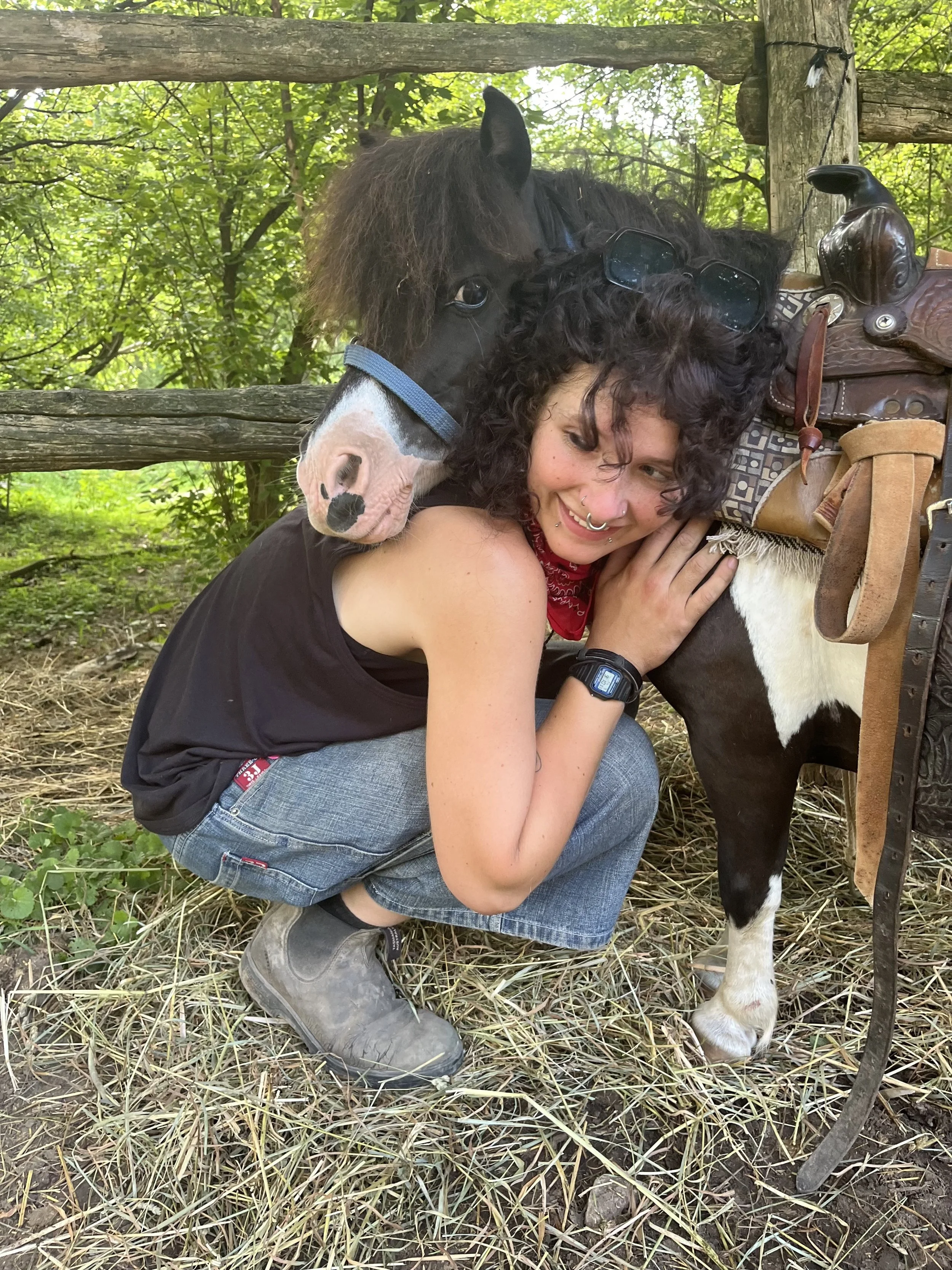 Young woman with red curly hair and septum piercing, wearing a black beanie and gray zip-up jacket, standing outdoors with a playful expression including sticking out her tongue. In the background, there is a woman in a helmet sitting on a chair and a person riding a horse with white fur and saddle.