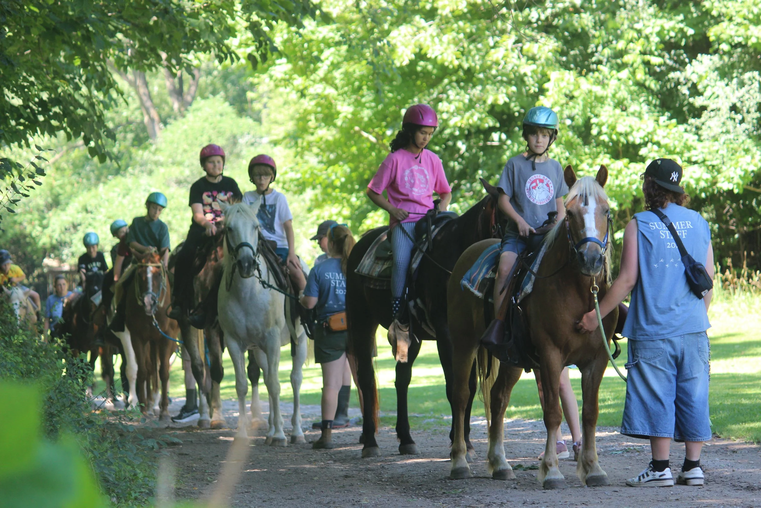 Children riding horses in a line through a wooded area, guided by adult instructors.