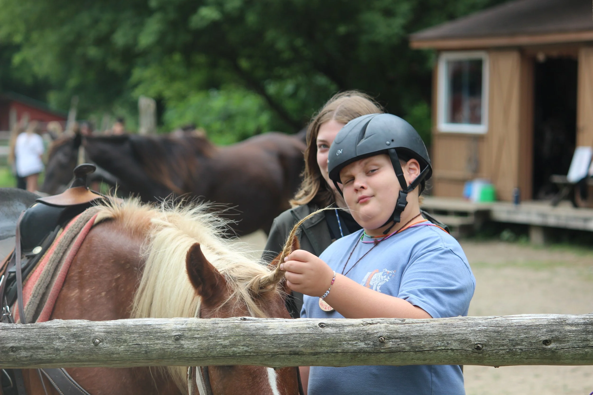 A boy wearing a helmet is petting a brown horse with a blonde mane, with a woman behind him, at an outdoor farm or stable area.