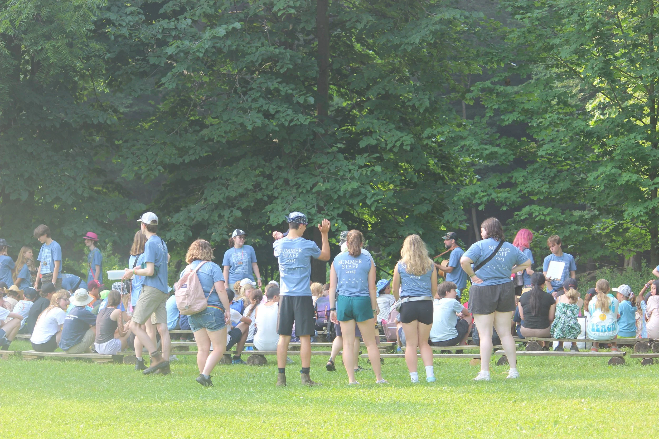 Group of people outdoors sitting on grass and benches, some standing, many wearing blue "Summer Staff" t-shirts, surrounded by green trees.