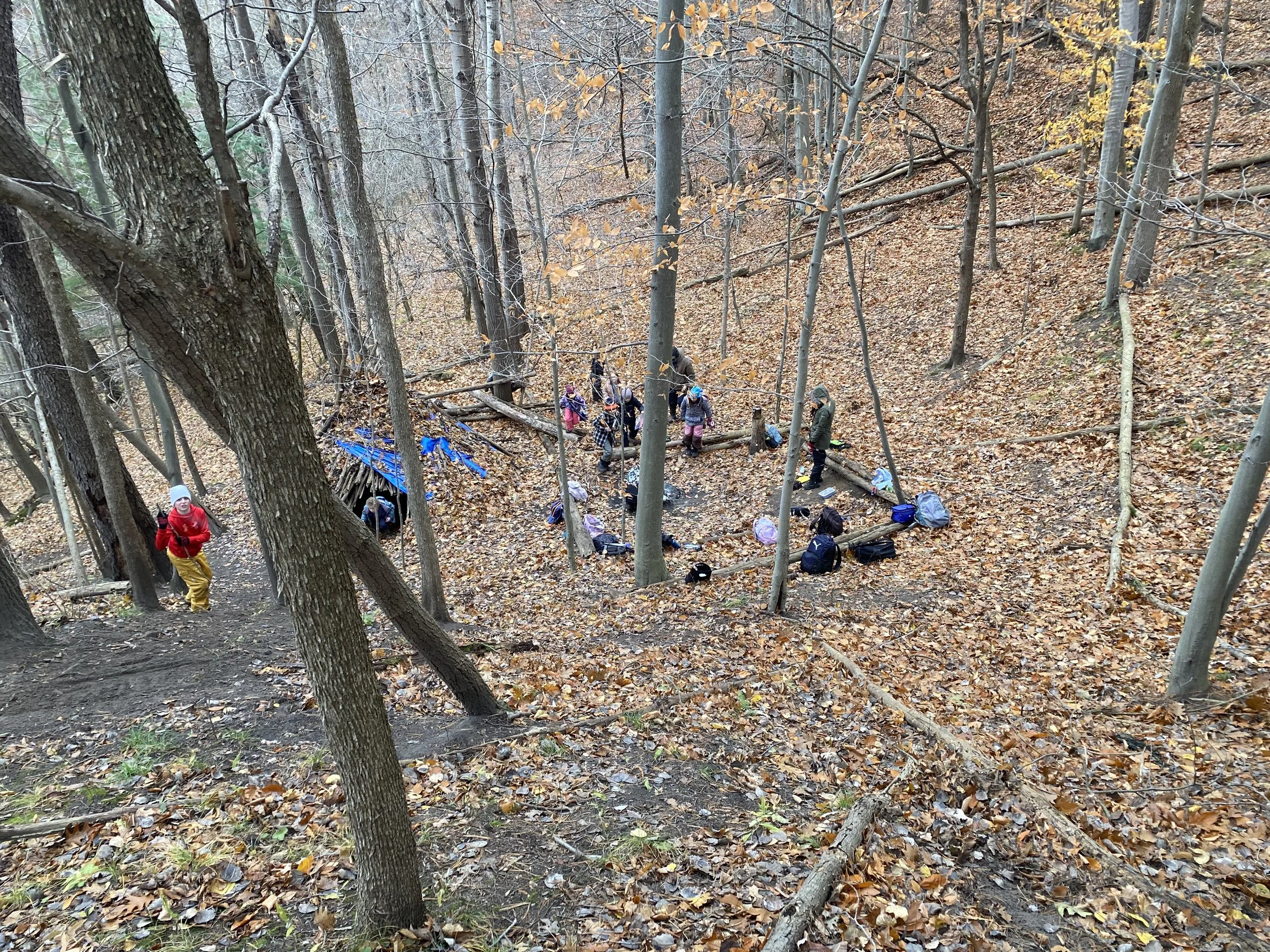 A group of children and adults in a wooded area during fall, some sitting around a makeshift camp with a blue tarp, backpacks, and supplies, with fallen leaves on the ground.