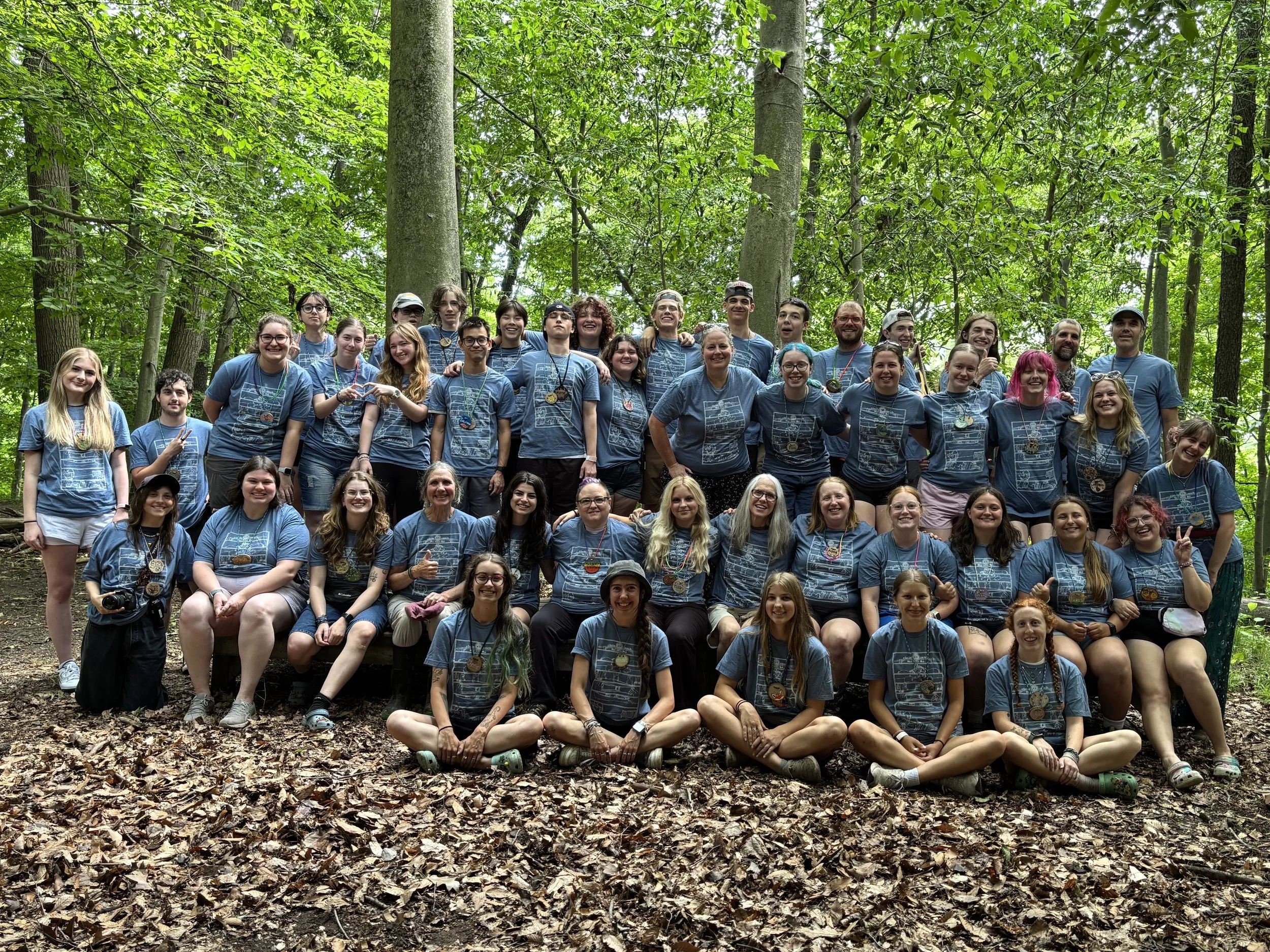 A large group of people in matching blue T-shirts posing for a photo in a forested area with green trees and brown leaves on the ground.