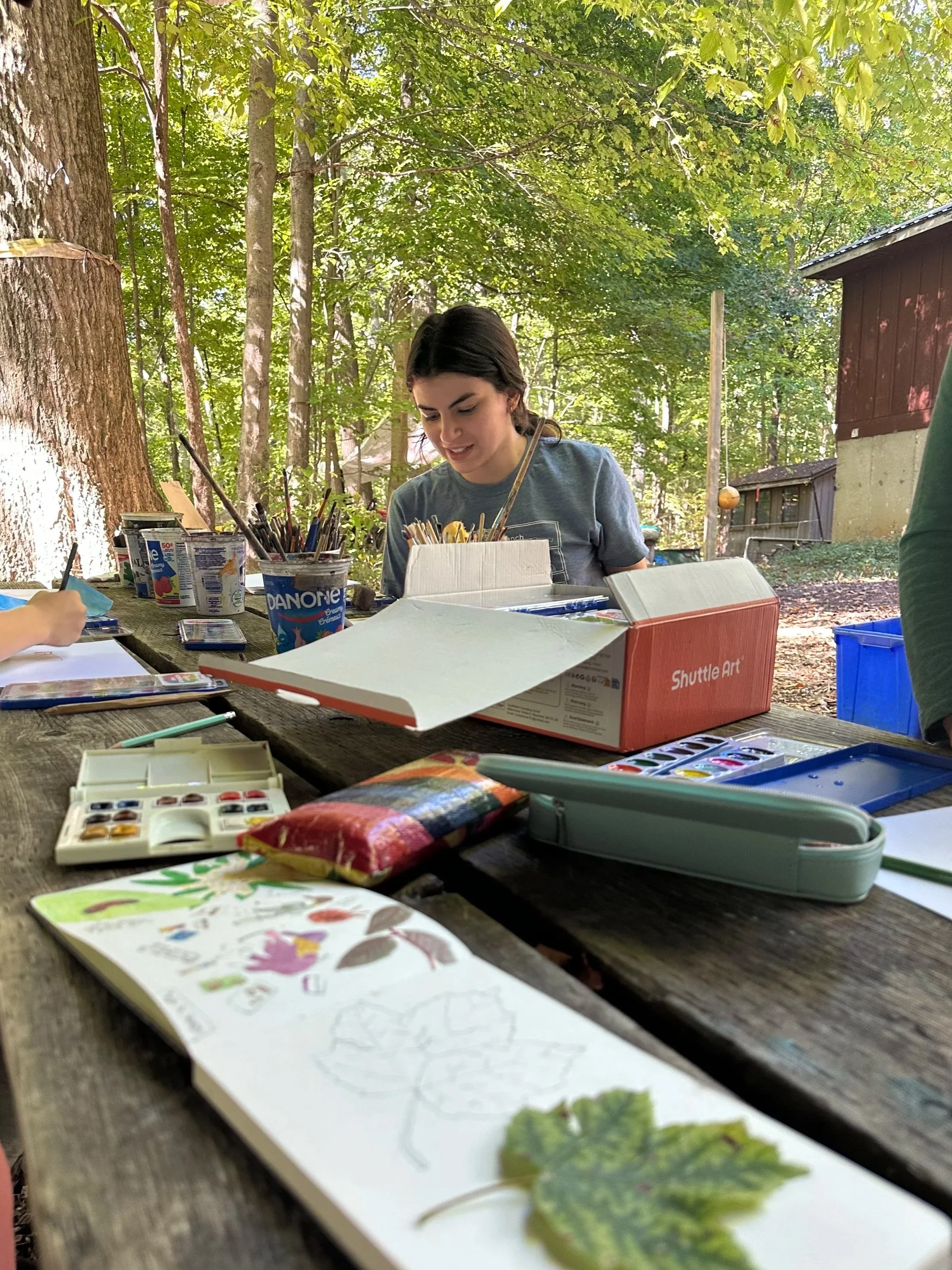 A woman sitting at a picnic table outdoors in a forest, engaged in an art activity with various watercolor supplies, paper, and nature leaves on the table.