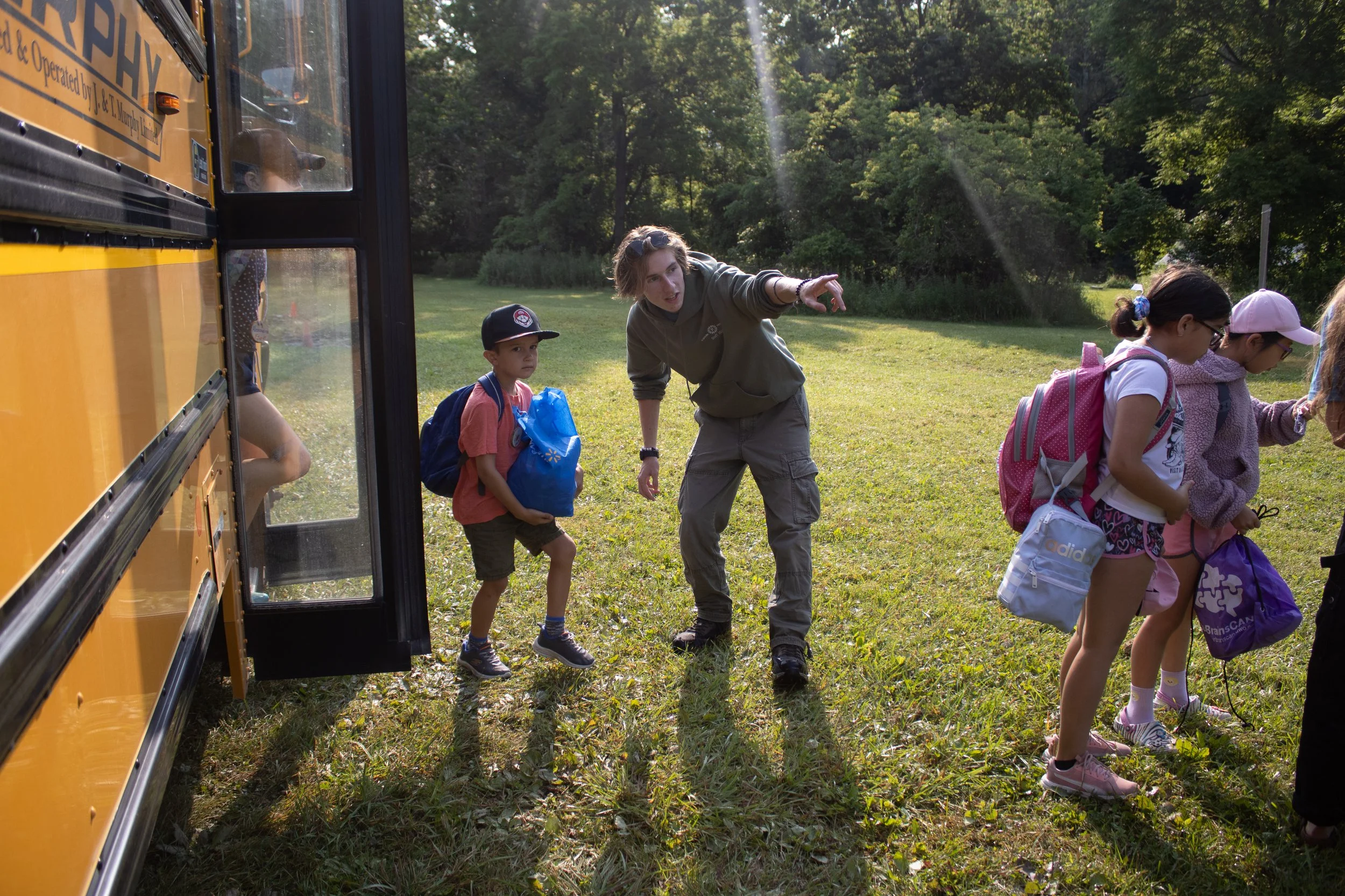 A girl with a backpack talks to a younger boy with a backpack near a yellow school bus in a grassy field with trees in the background.